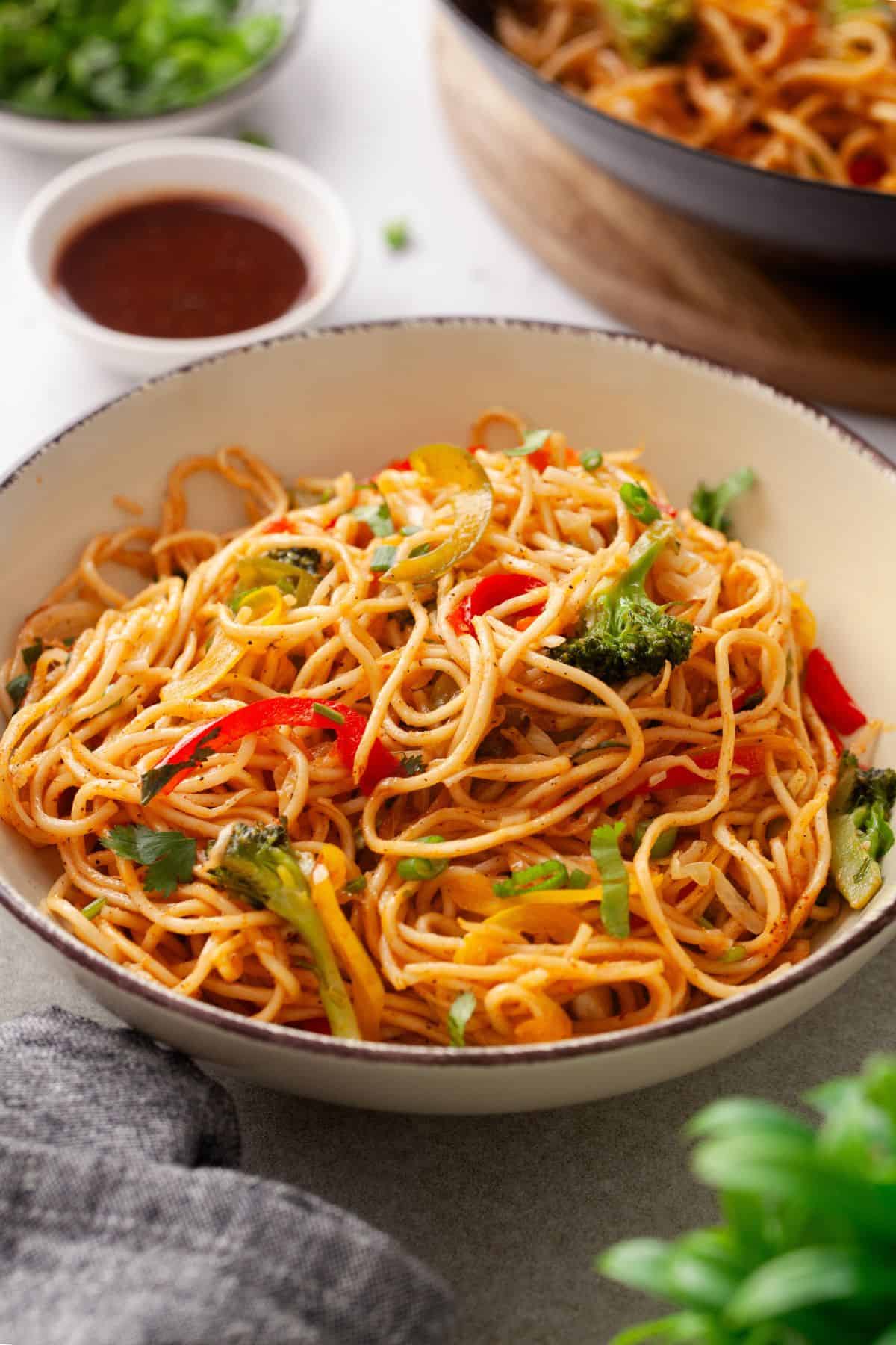 Vegetable hakka noodles are served in a cream colored bowl with sauce and herbs in background.