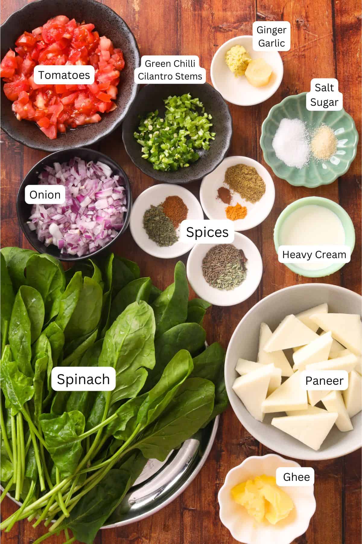 A flat lay of ingredients for palak paneer on a wooden surface: chopped tomatoes, onion, green chili and cilantro stems, ginger garlic, assorted spices, salt, sugar, heavy cream, ghee, cubes of paneer, and fresh spinach leaves.