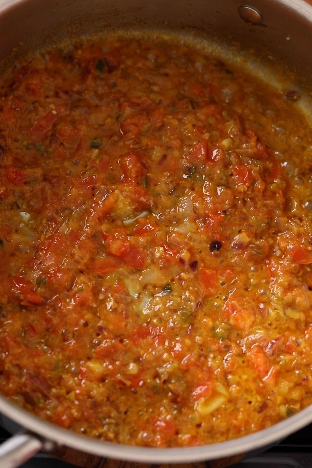 A close-up of a pot filled with simmering tomato-based sauce, showing chunks of tomatoes, onions, spices, and herbs in a thick, orange-red mixture