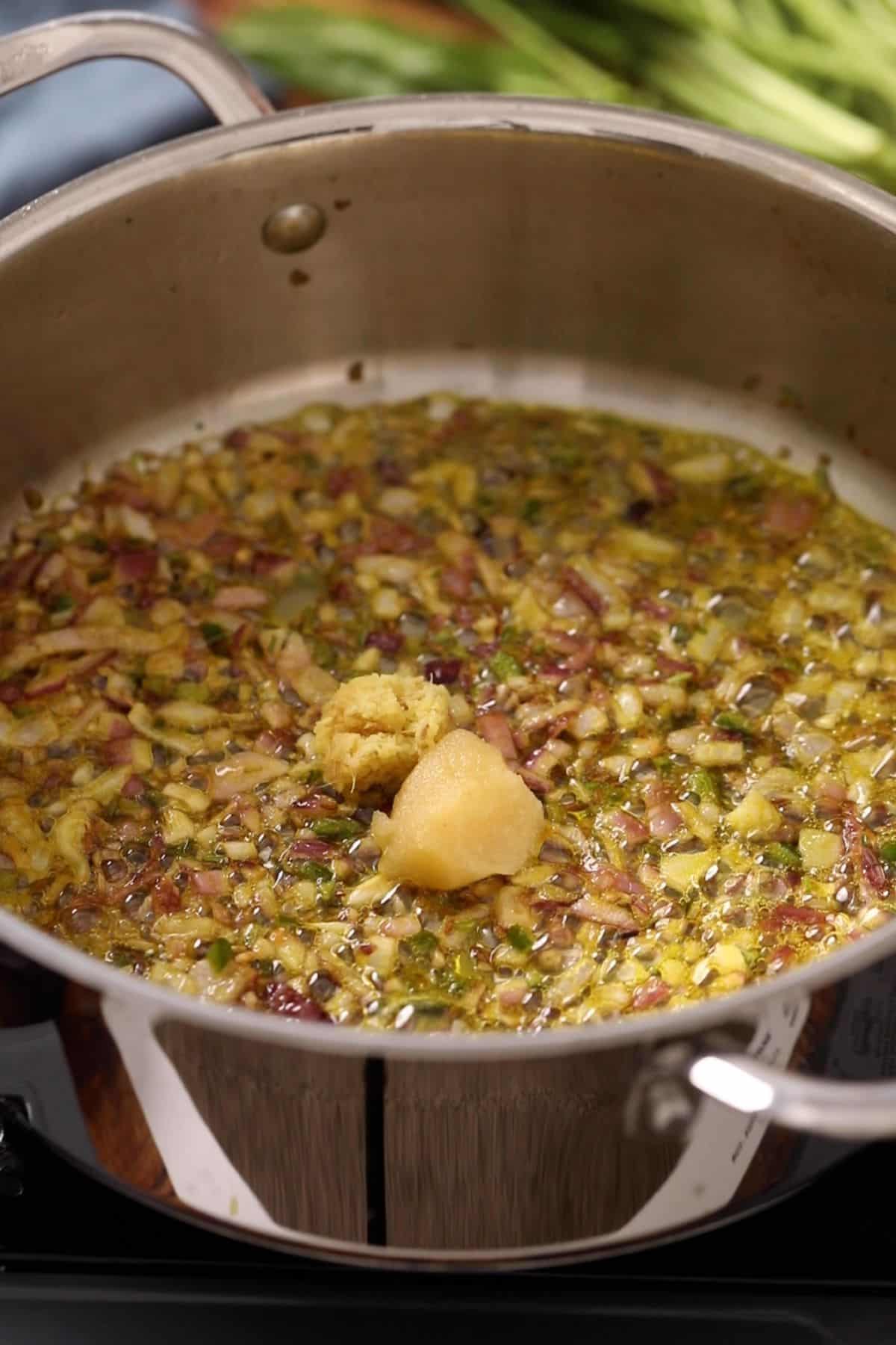 Chopped onions, green herbs, and a dollop of ginger-garlic paste are sautéing in oil in a stainless steel pan on a stovetop.