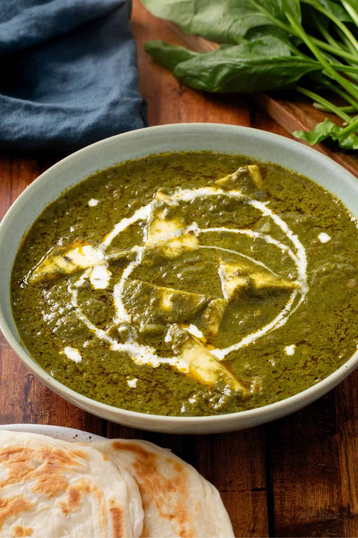 A bowl of palak paneer garnished with cream. Beside the bowl are flatbreads and fresh spinach leaves on a wooden surface.