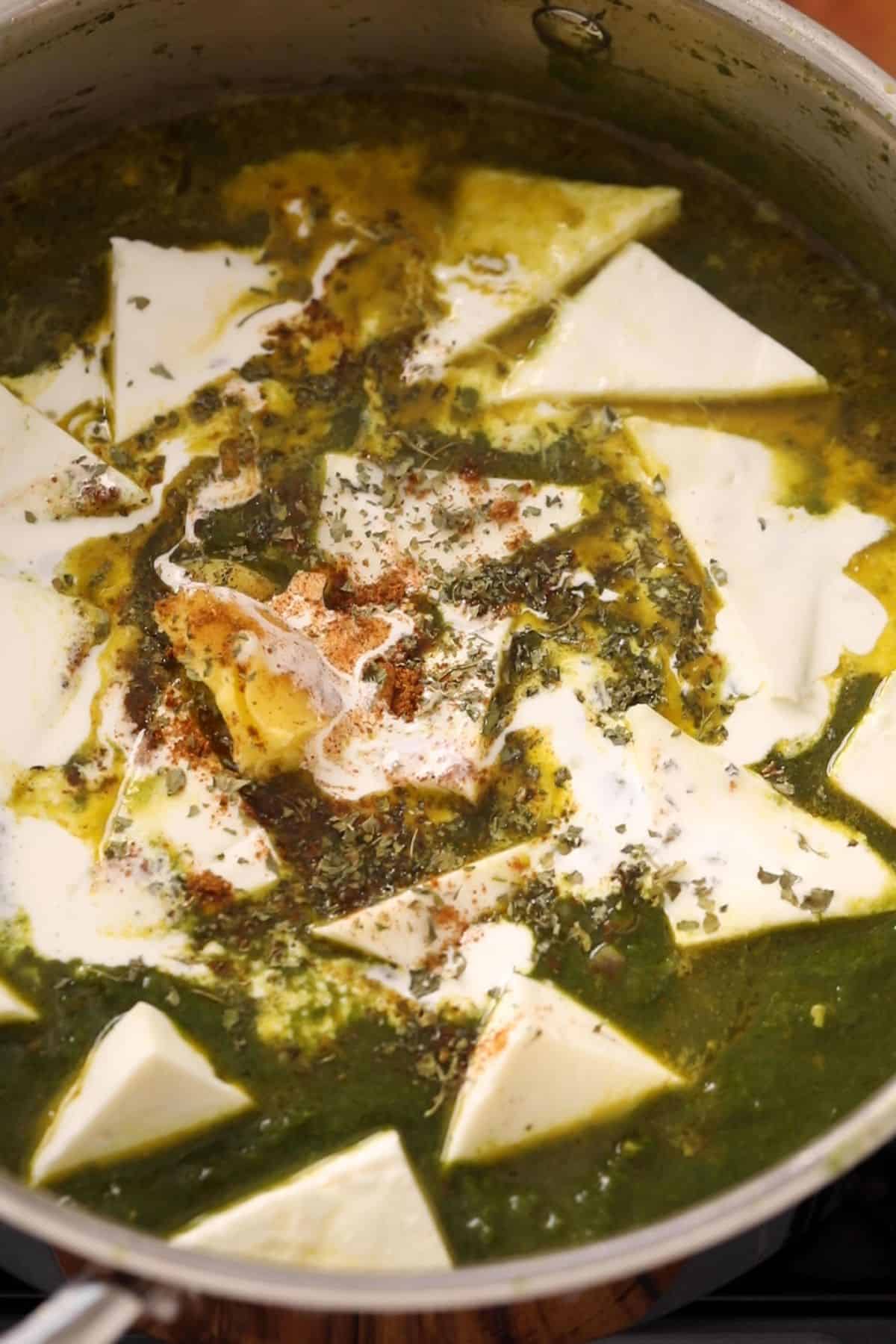 A close-up of a pot filled with palak paneer, featuring white paneer cubes in a creamy spinach gravy, garnished with cream, butter, and sprinkled spices.