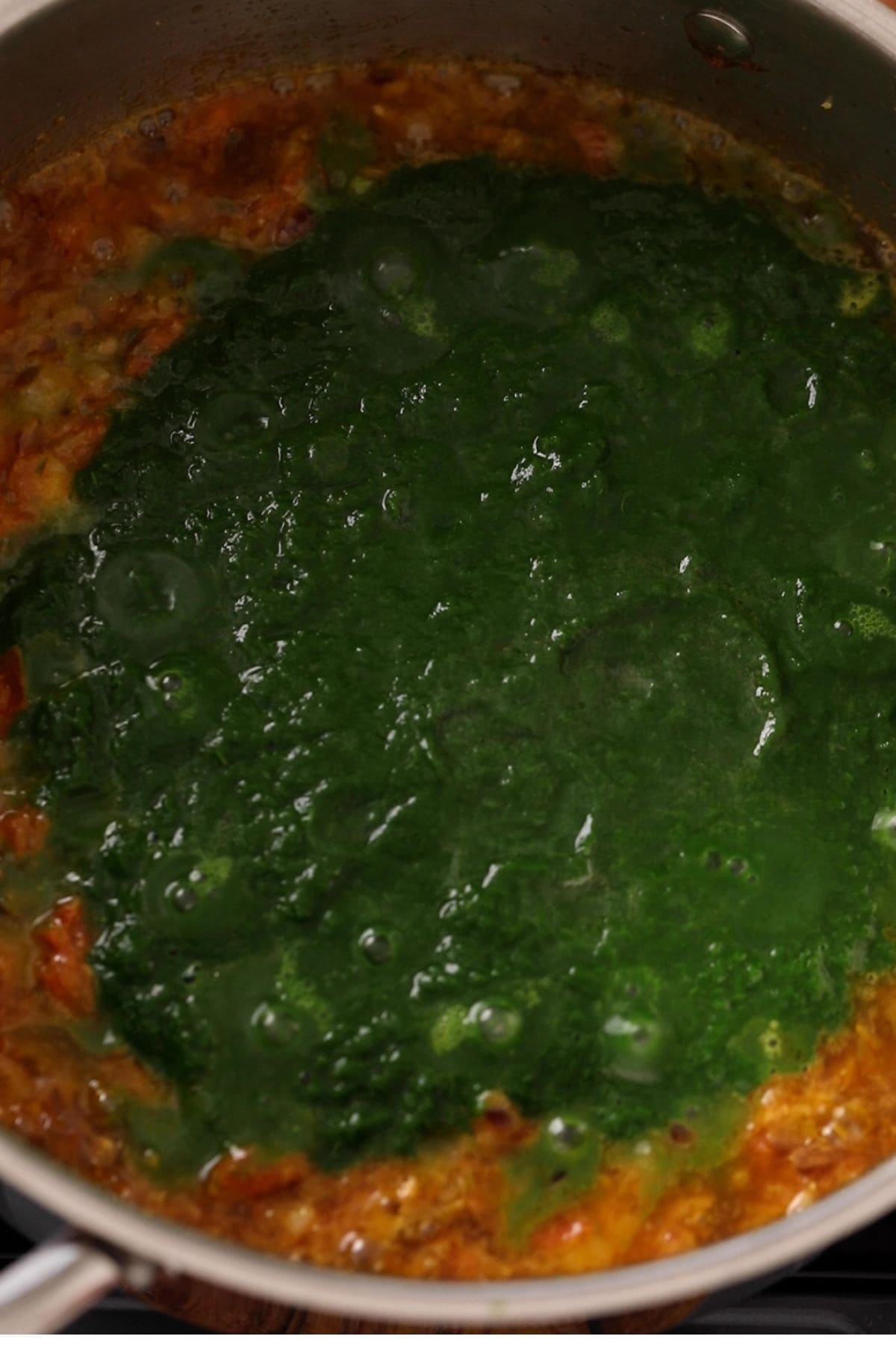 A pot on a stove contains a simmering mixture of green spinach puree on top of a reddish-brown sauce, likely the preparation of a traditional Indian dish such as saag or palak paneer.