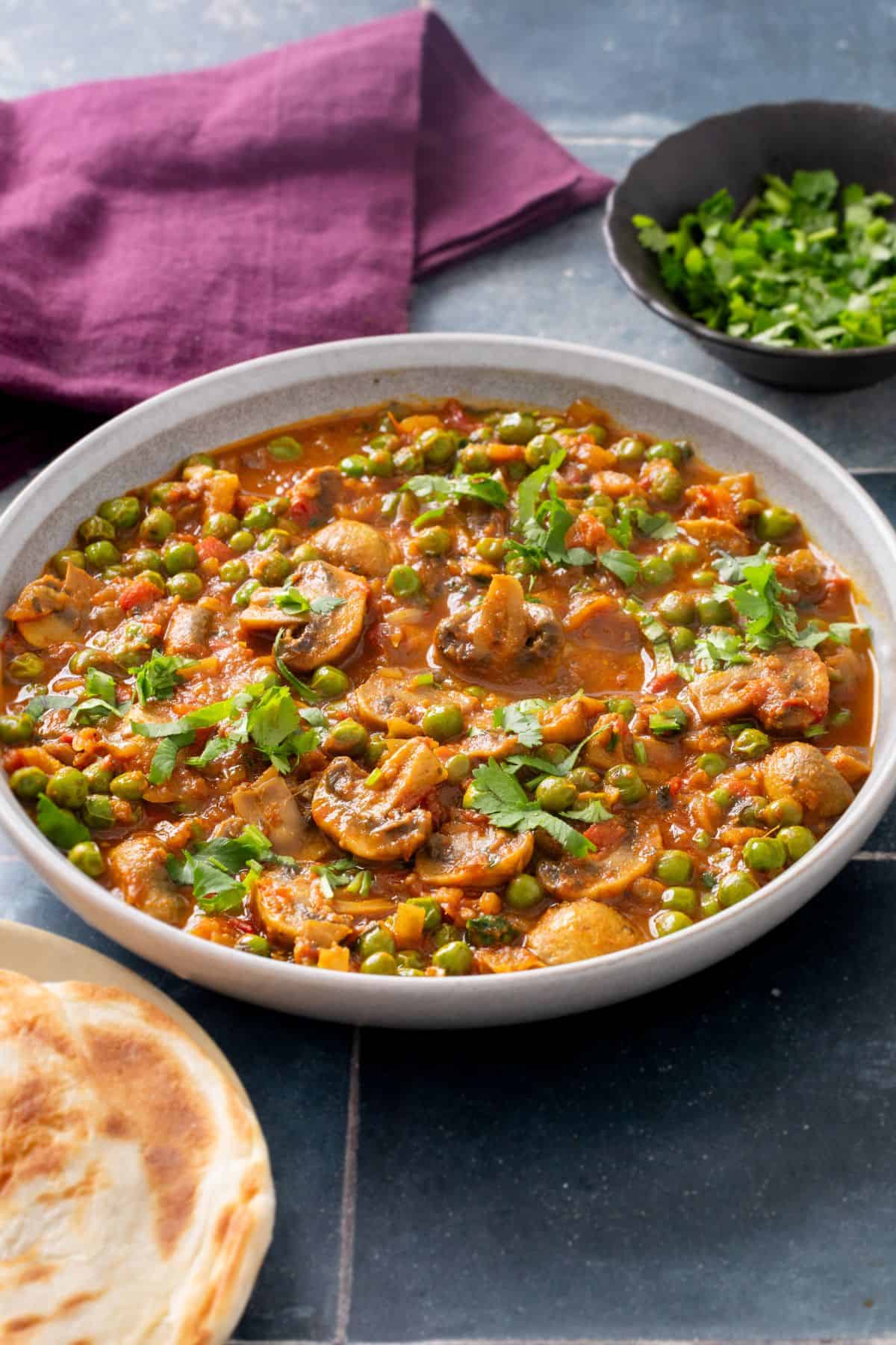 Mushroom Matar Masala is served in a grey bowl with flatbreads and cilantro placed around it.