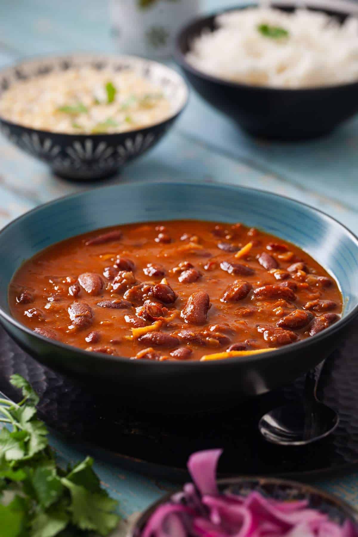 Rajma curry is served in a blue bowl with raita and steamed white rice placed in the background.