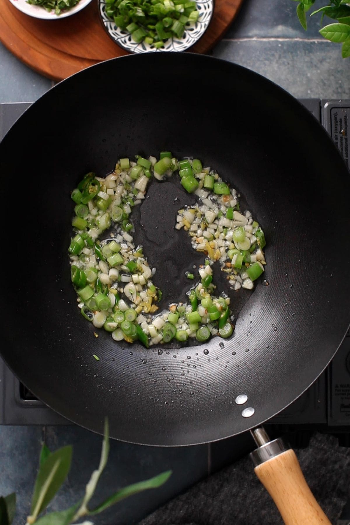 Chopped green onions, garlic, and ginger are being sautéed in a black wok on a stovetop, with bowls of additional chopped green onions visible in the background.