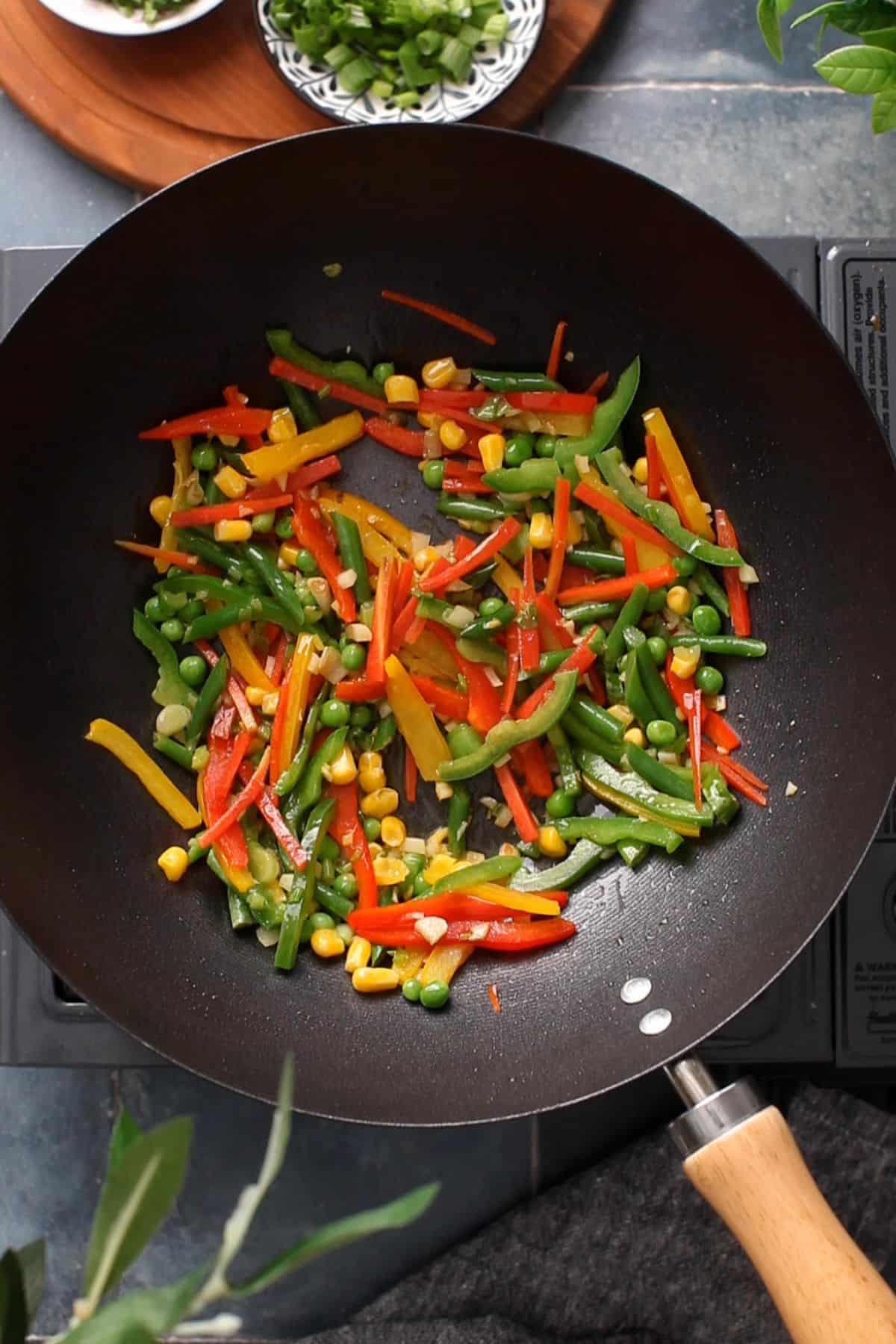 Colorful sliced bell peppers, green beans, corn, and peas are being stir-fried in a black wok on a stovetop, with some fresh herbs and chopped green onions visible in small bowls nearby.