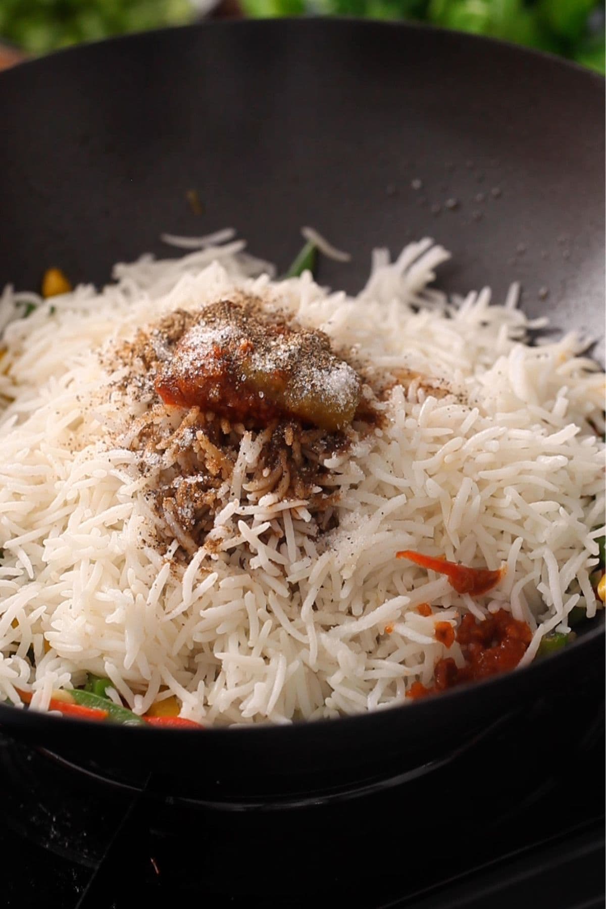 A close-up of cooked white rice in a pan, topped with sauces, spices, and black pepper, with some visible vegetables underneath.