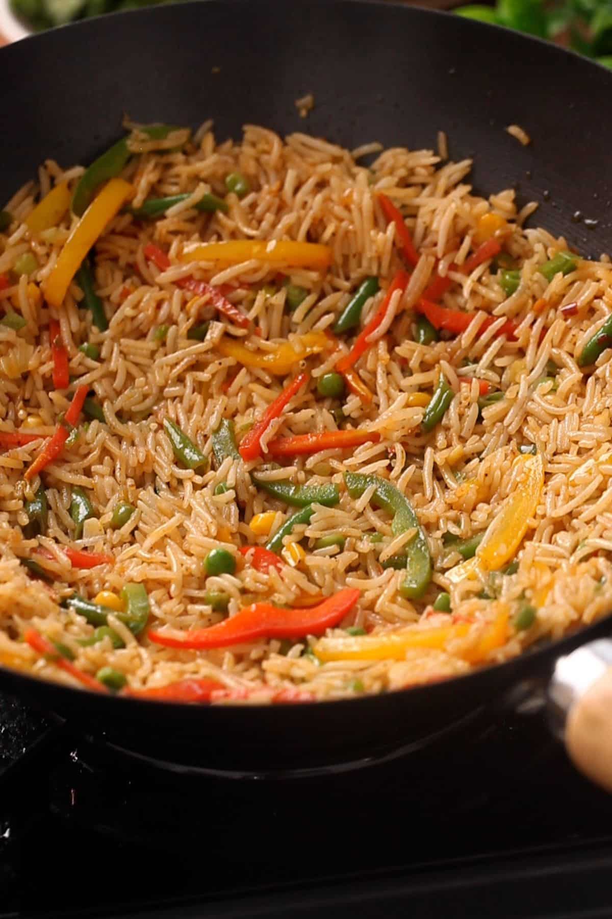 A wok filled with fried rice mixed with colorful sliced bell peppers, green beans, and peas, being cooked on a stovetop.