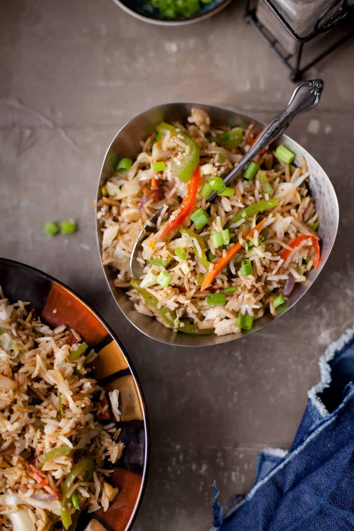 A triangular bowl of fried rice with vegetables, including red bell peppers and green onions, sits on a table next to a fork and a napkin. Another bowl of fried rice is partially visible nearby.