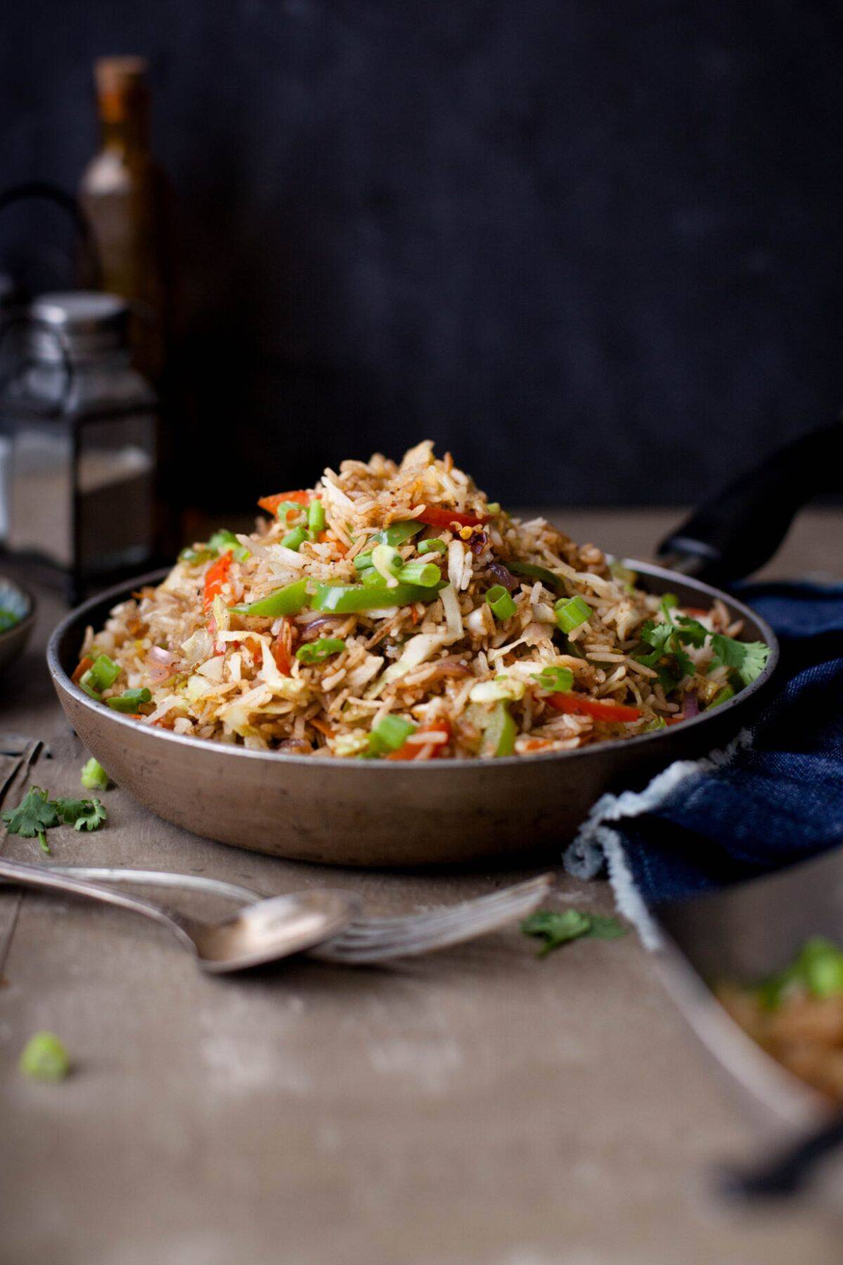 A rustic bowl filled with vegetable fried rice garnished with chopped green onions, placed on a wooden table with a fork and napkin nearby. The background is softly blurred, creating a cozy, inviting atmosphere.