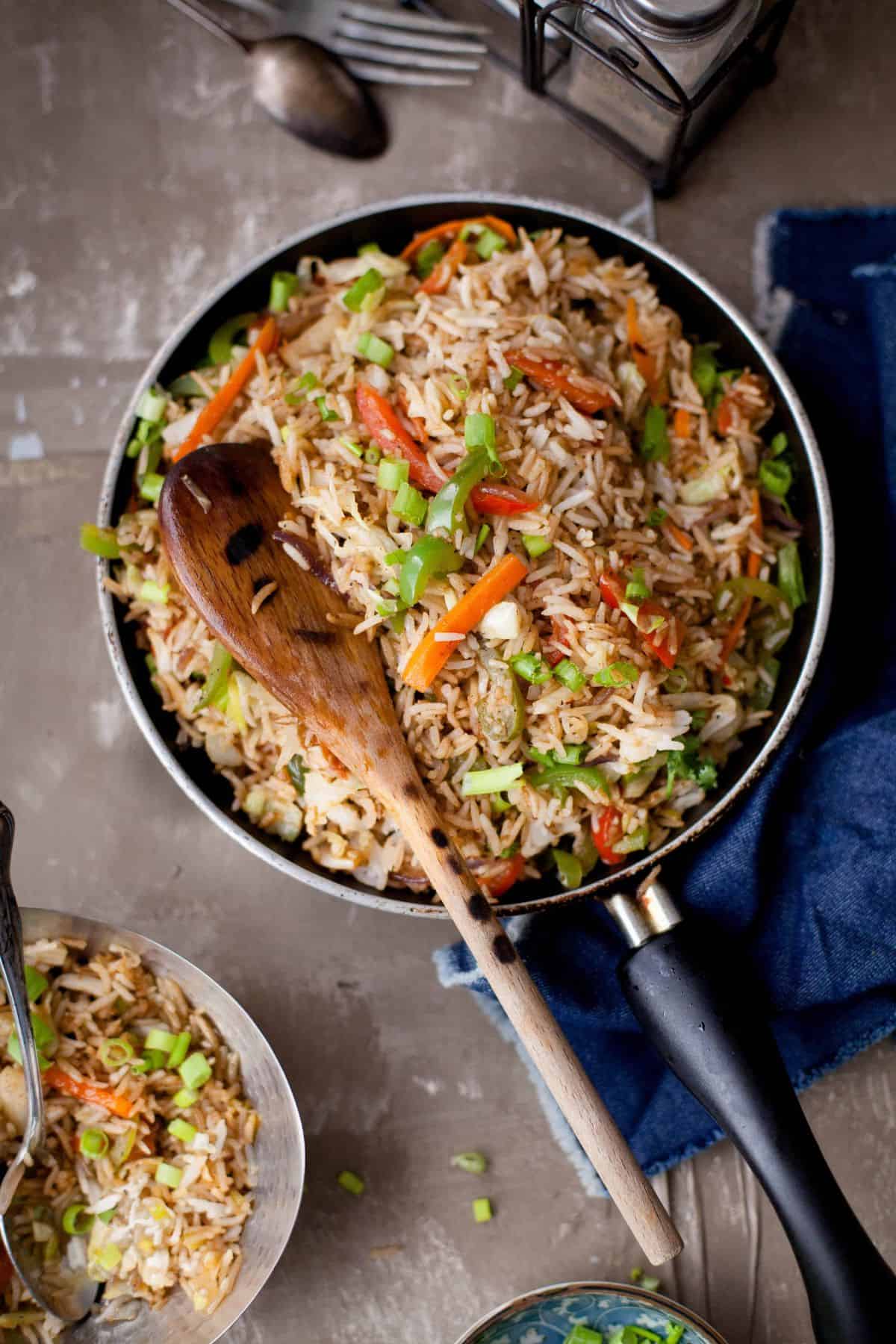 A pan of vegetable fried rice with carrots, peppers, and green onions, garnished with chopped scallions. A wooden spoon rests on the pan, and a fork and napkin are nearby on the table.