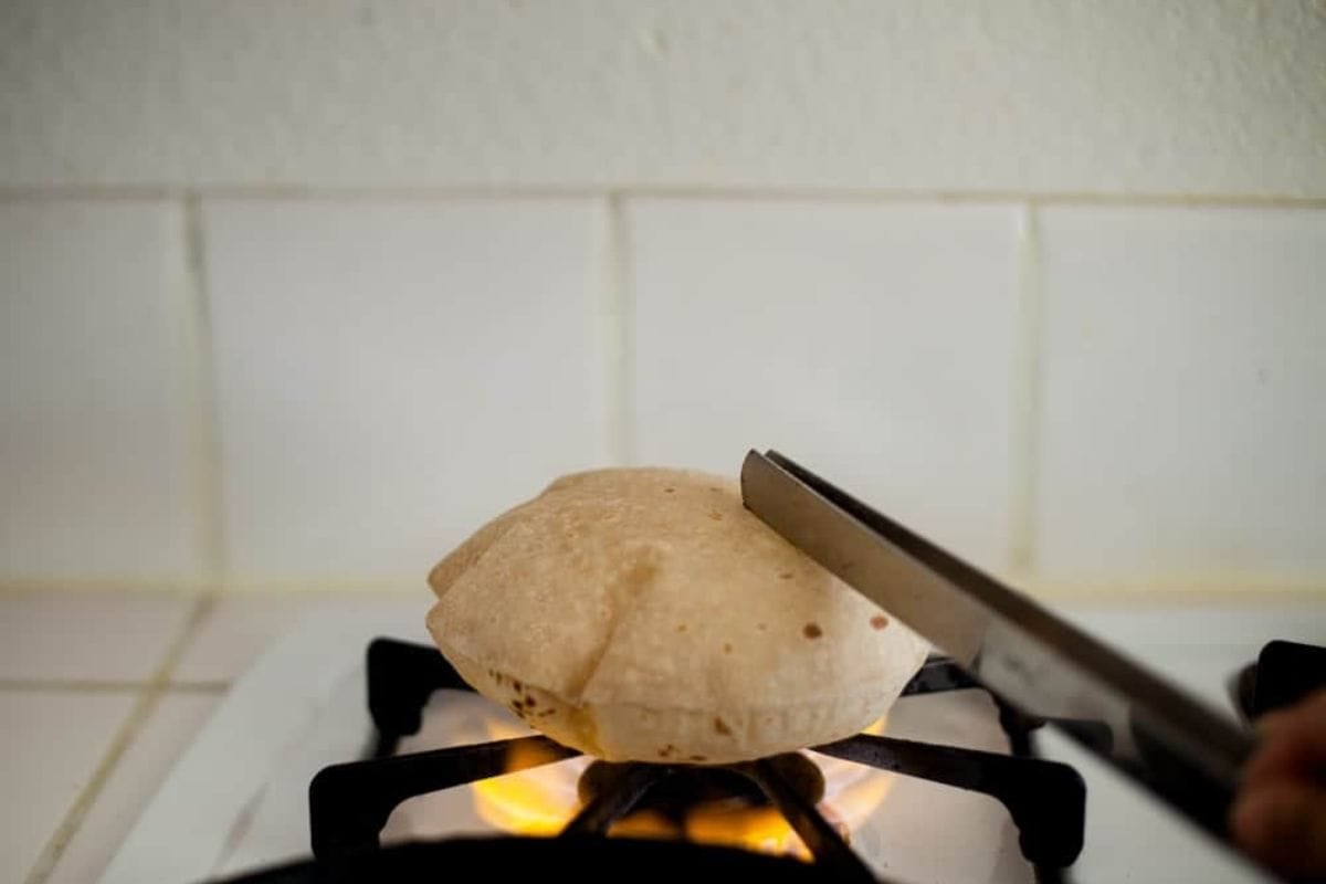 Image shows soft puffy roti on open flame that is being pressed with steel tongs.