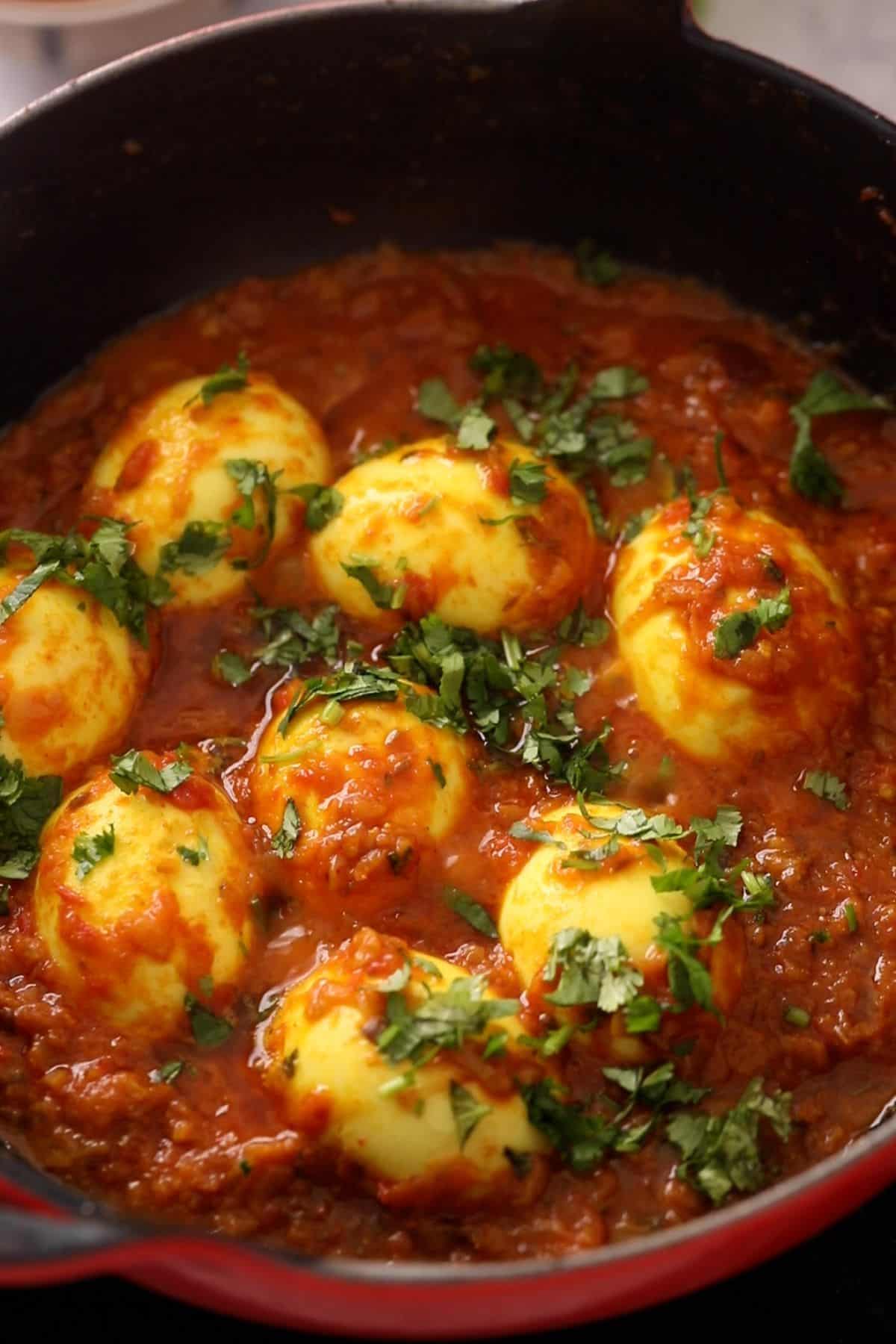 Hard-boiled eggs simmer in a rich, spiced tomato curry sauce, garnished with chopped cilantro, served in a black cast iron pan.