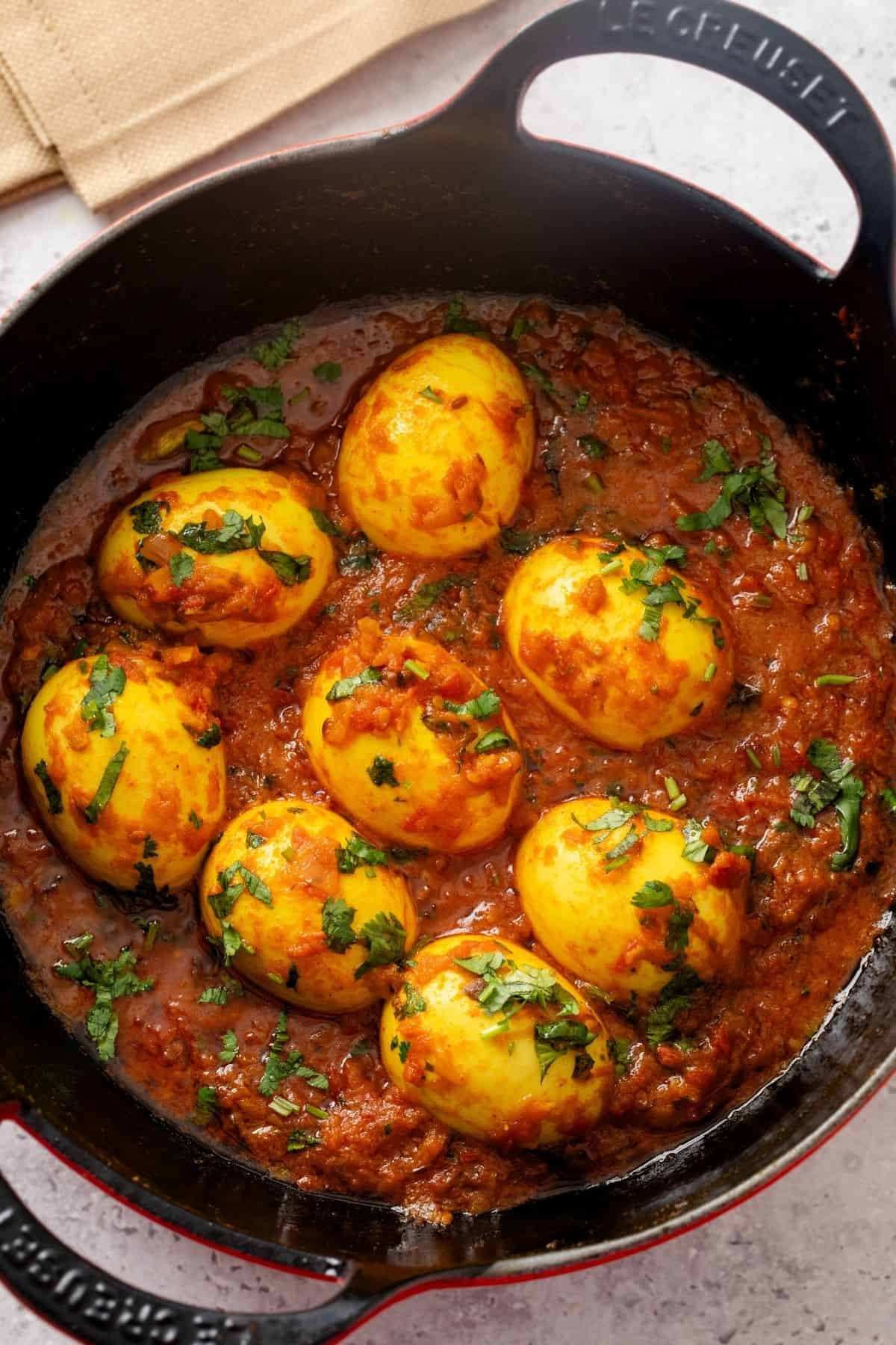 Hard-boiled eggs simmer in a rich, spiced tomato curry sauce, garnished with chopped cilantro, served in a black cast iron pan.