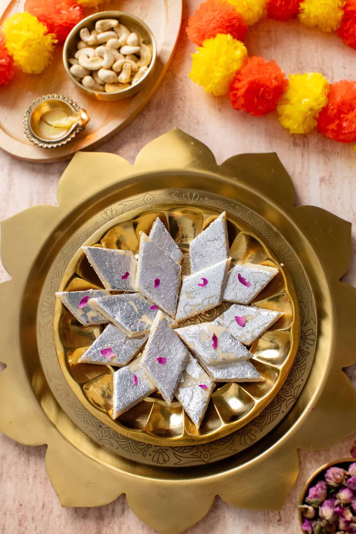 Kaju Katli placed in a lotus shaped platter with a lit diya and marigold garland.