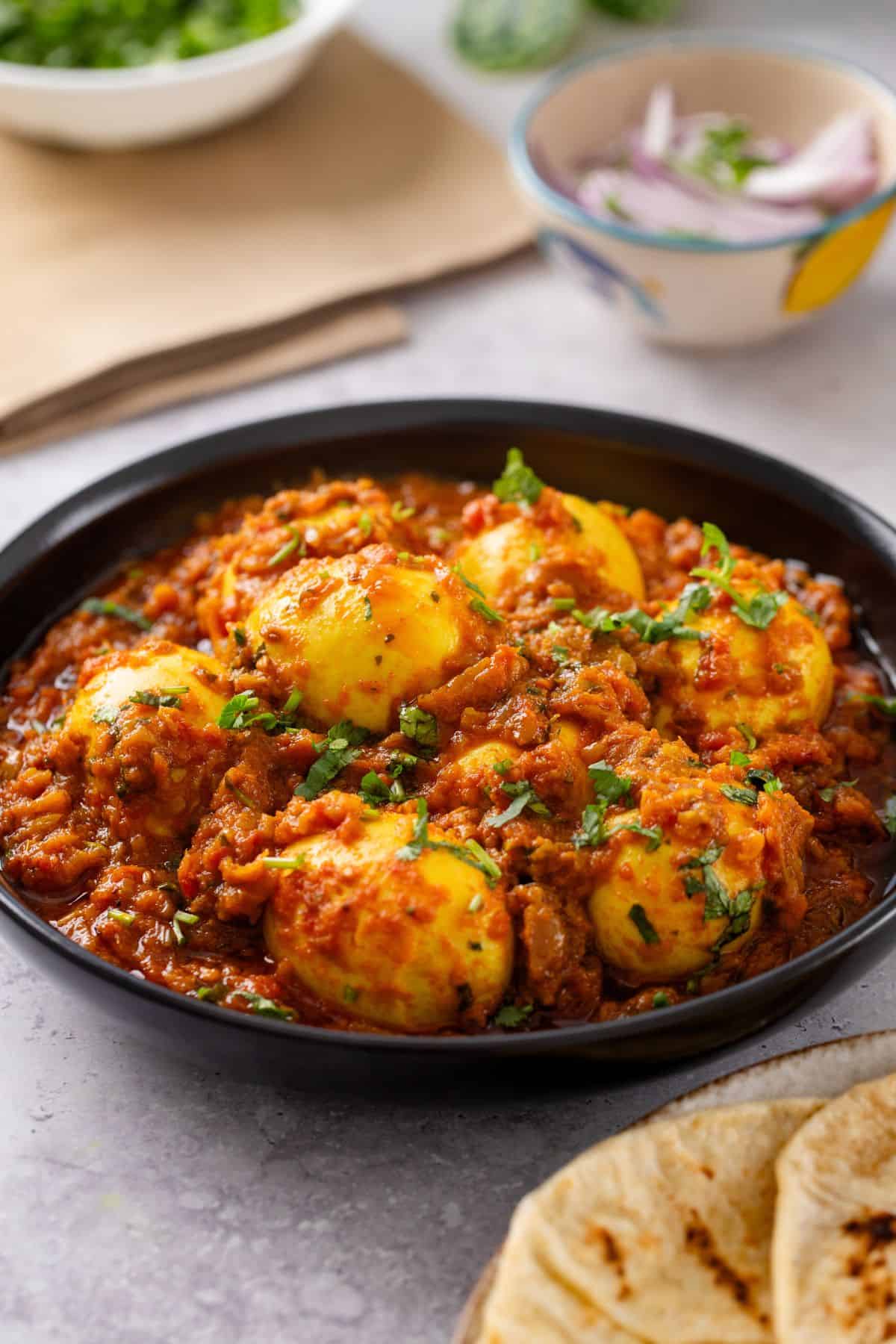 Punjabi anda curry is served in a black dish with sliced onions and roti around it.