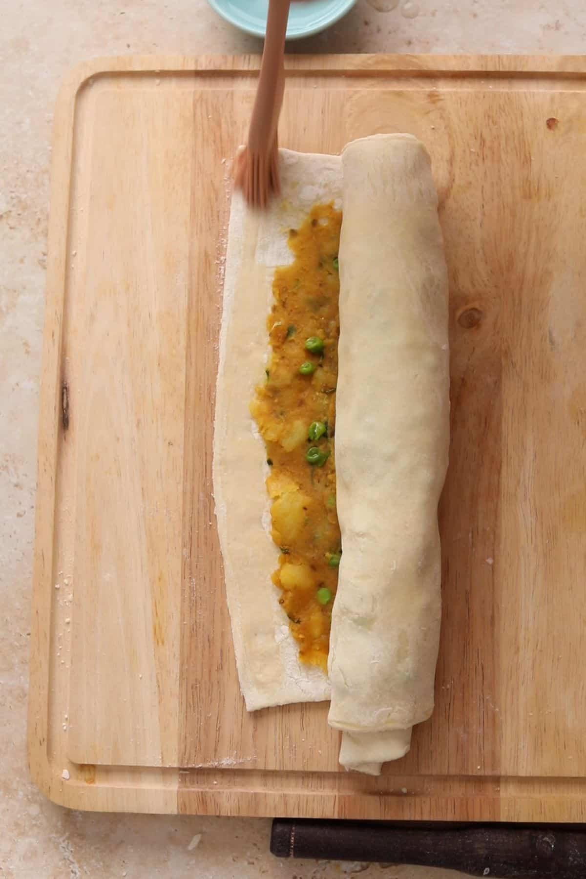 Rolling up pastry spread with a potato and green pea filling on a wooden cutting board.
