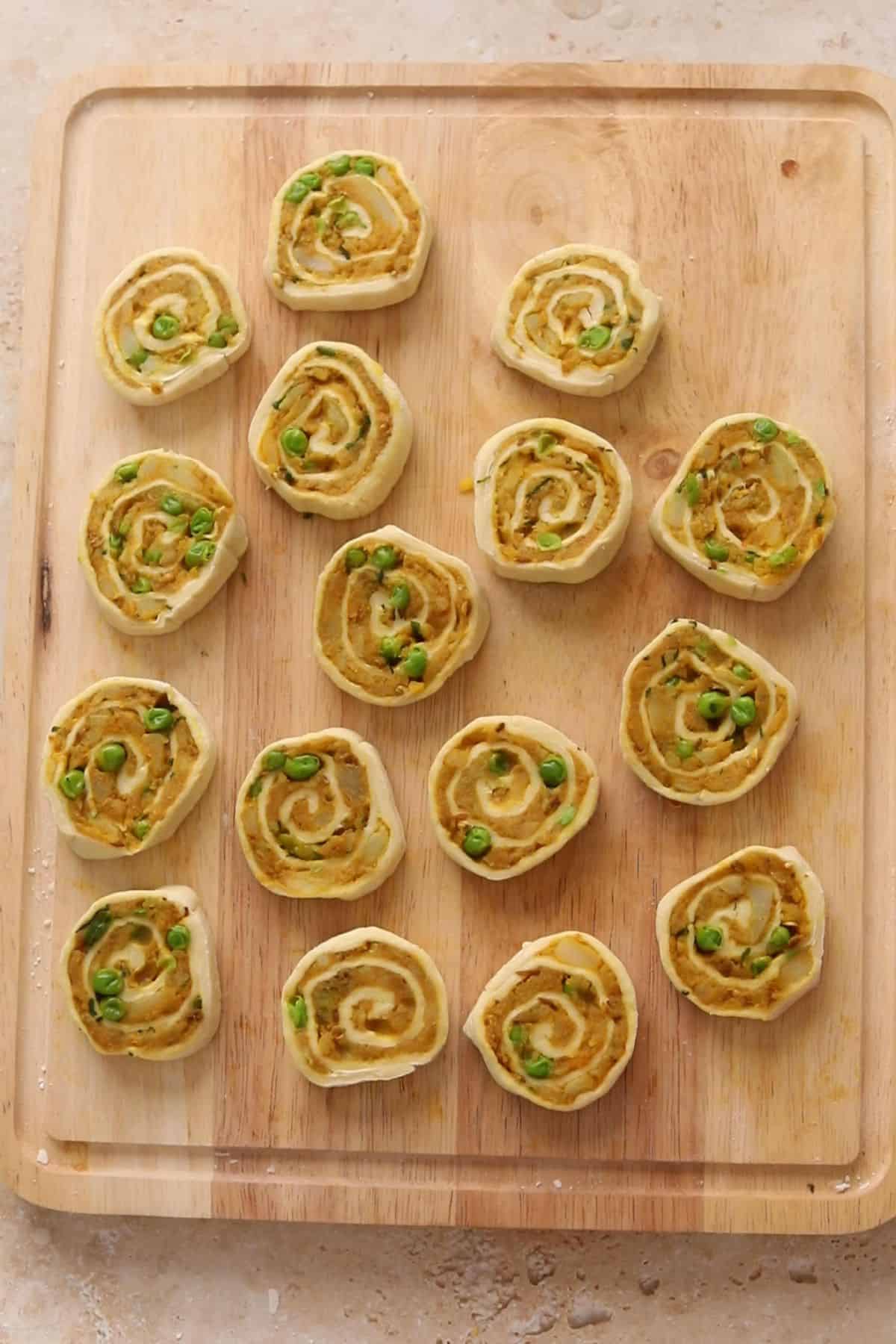 A wooden board with sixteen uncooked samosa pinwheels, arranged in a loose grid pattern.