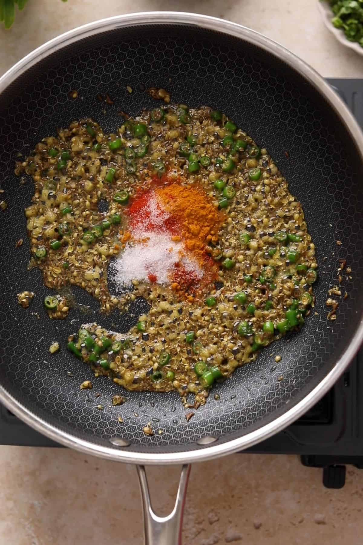 Whole and ground spices are frying in ghee along with aromatics in a large black skillet.