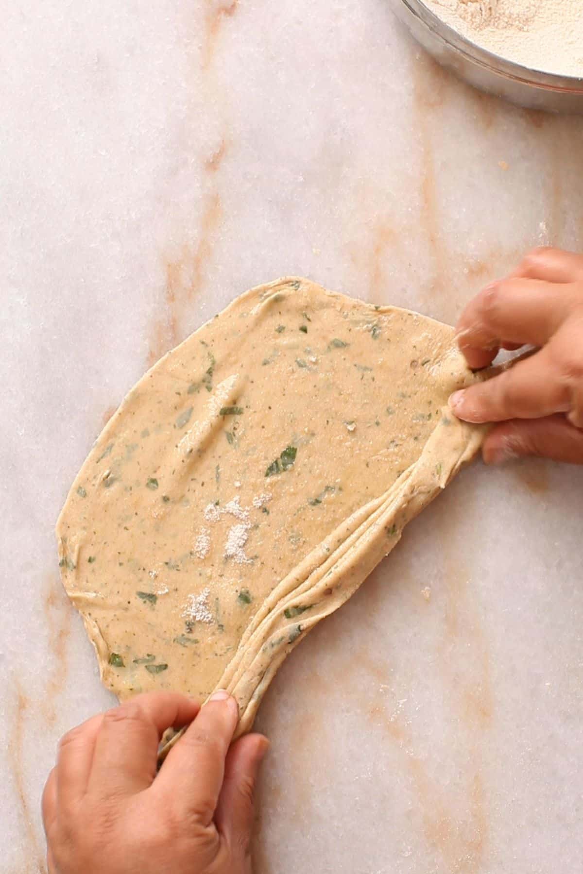 Stretching and folding dough like pleats with herbs on a light marble surface. A bowl of flour is partially visible in the upper right corner.