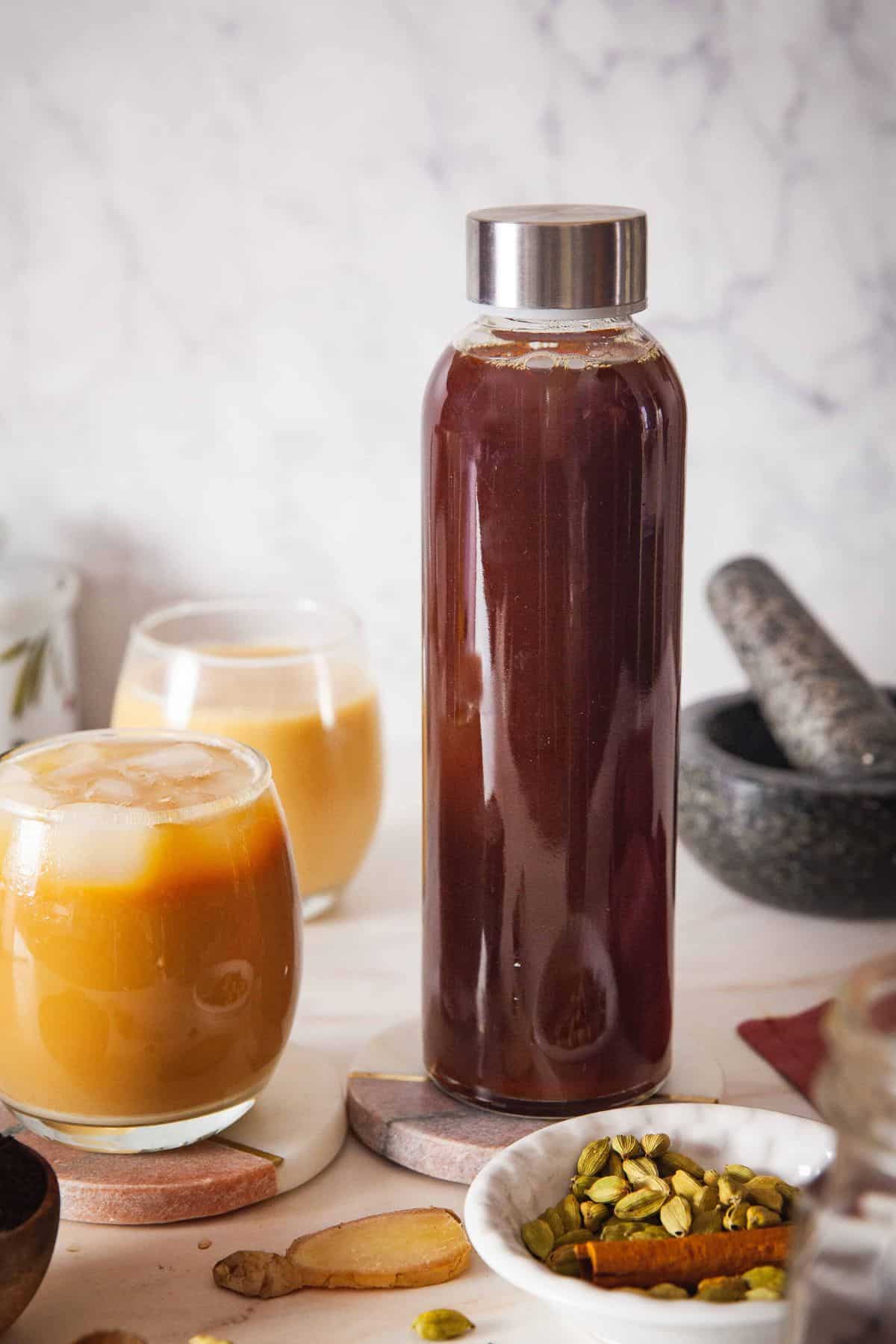DIY Chai Concentrate in a tall glass bottle alongside whole spices and a cup of ice chai on the side.