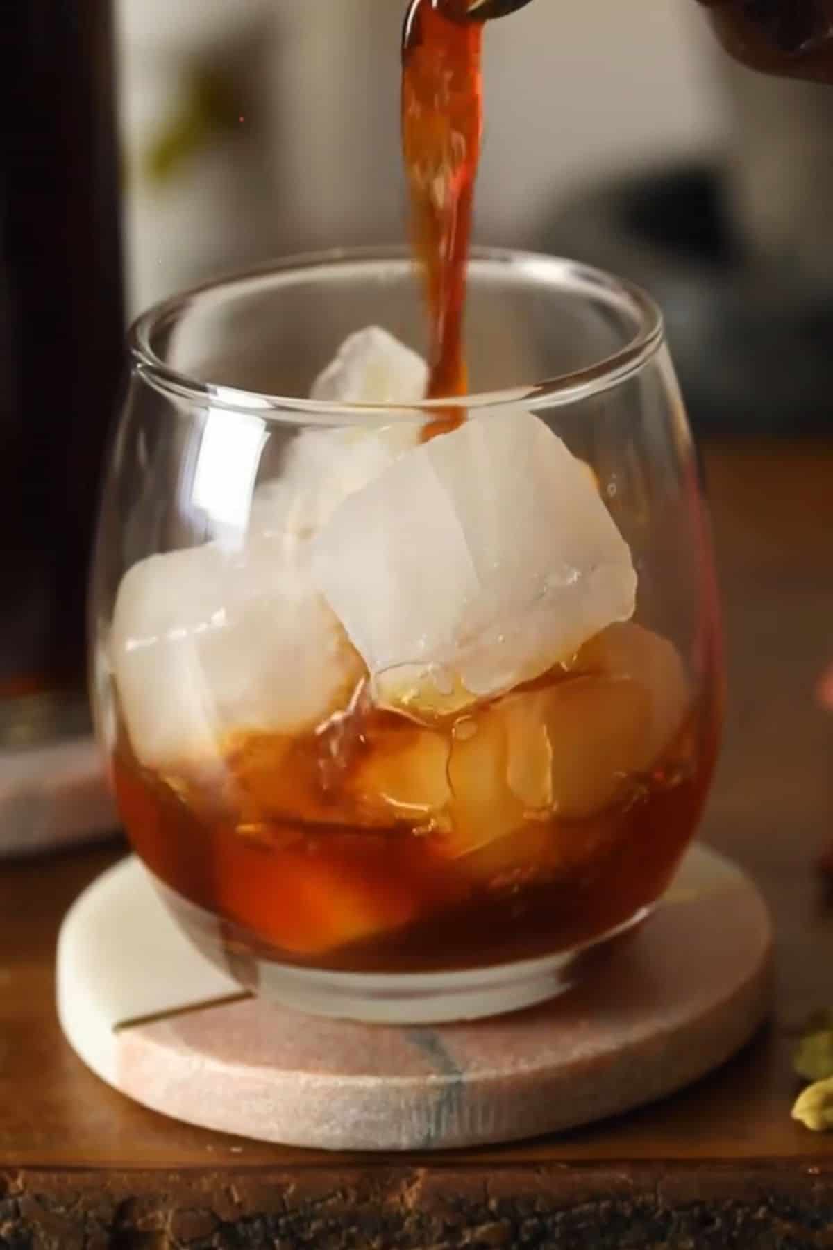 Chai concentrate is being poured over ice cubes in a glass cup.