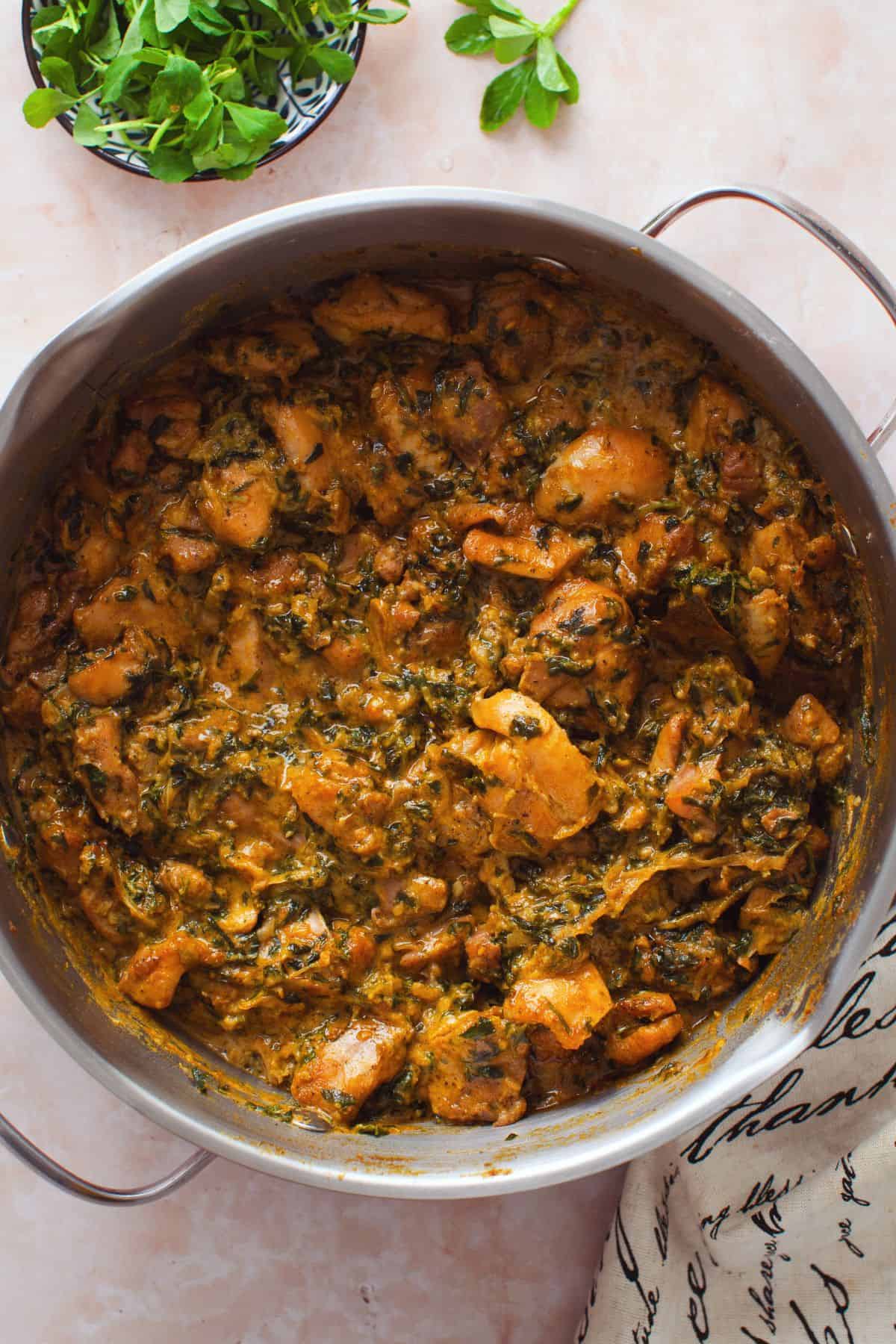 Cooked chicken methi curry in a wide steel cooking pot with fresh methi on the side.