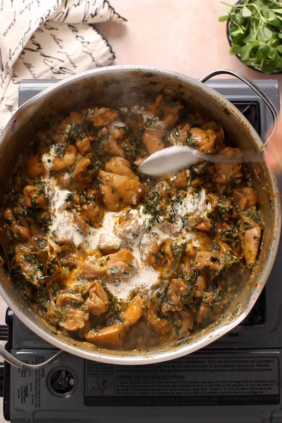 Heavy cream is added using a steel spoon to methi chicken.