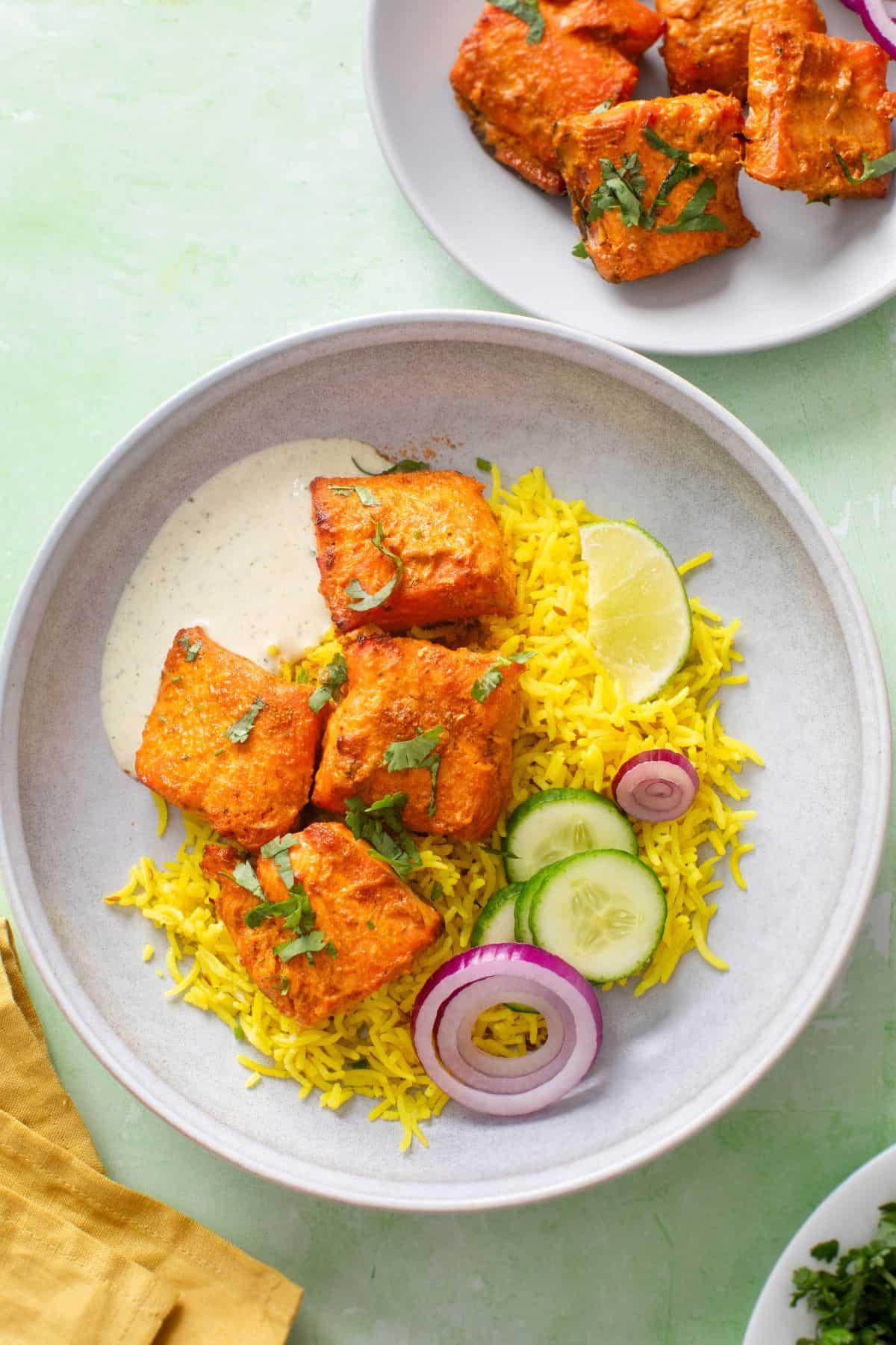 Salmon fish tikka is served with rice, cucumber and red onions along with thin yogurt in a grey bowl.