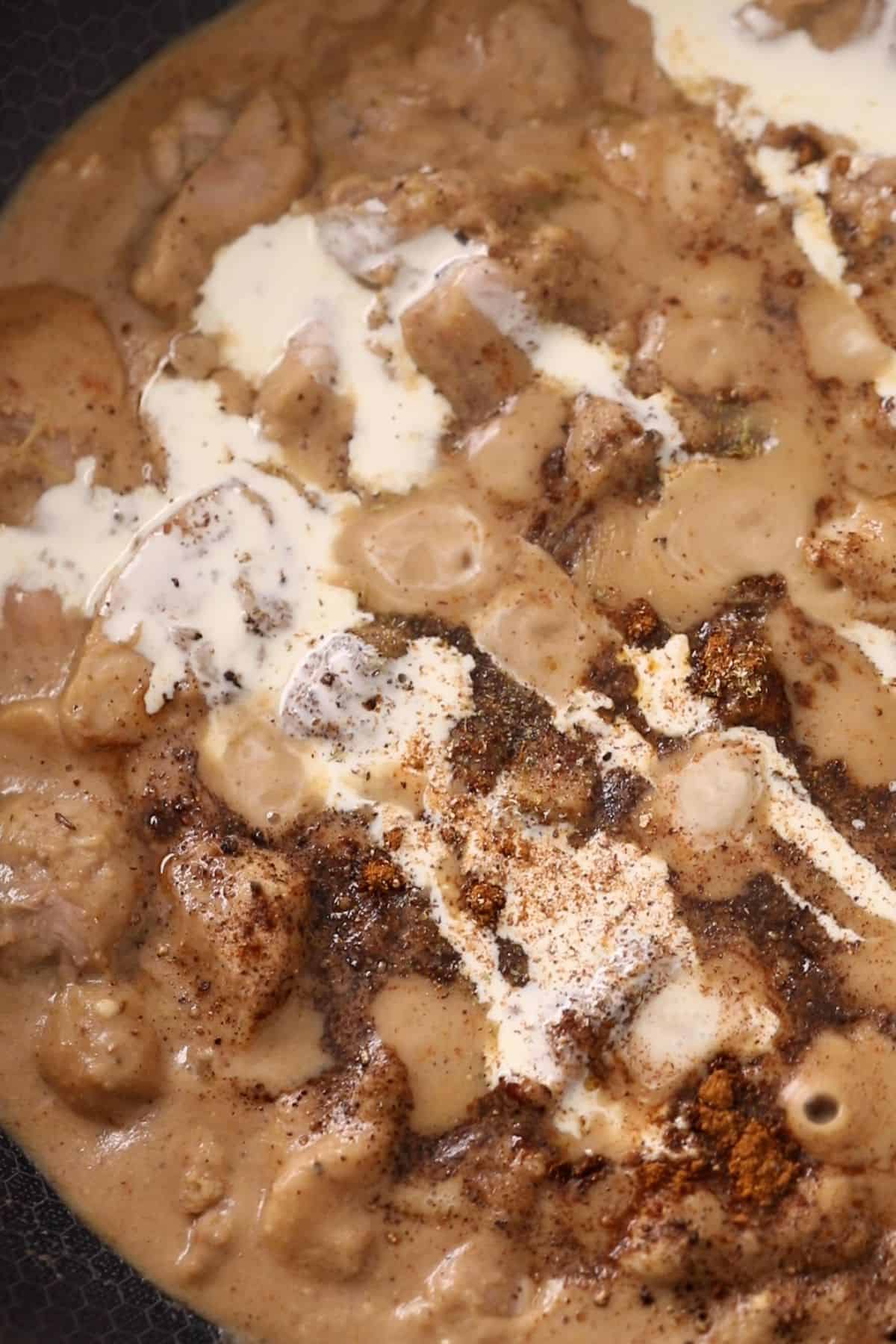 Chunks of chicken simmer in a creamy brown sauce, with visible swirls of cream and a sprinkle of spices on top, creating a rich and textured appearance.