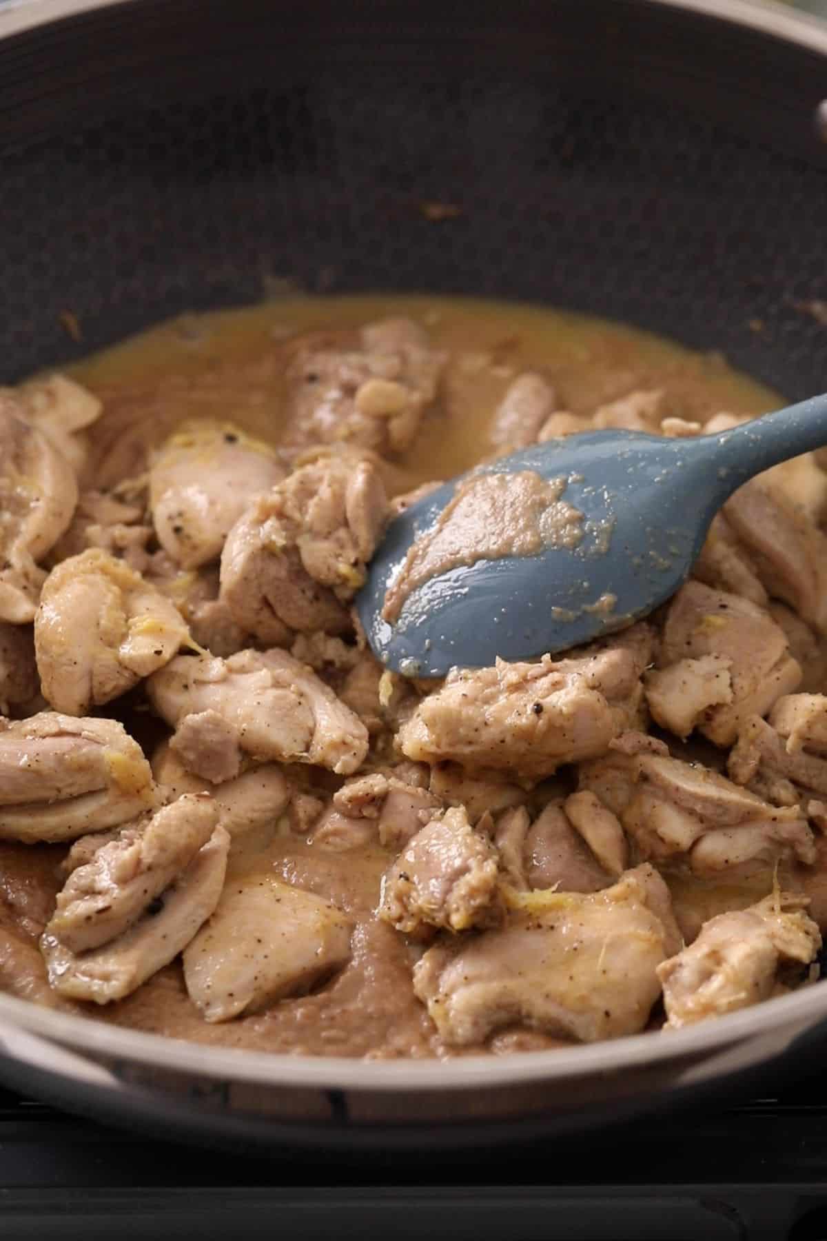 Chunks of seasoned chicken cooking in a black pan with sauce, being stirred with a blue spatula.