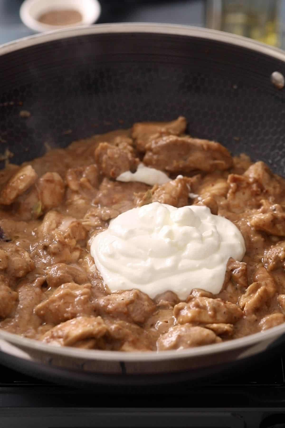 Chunks of cooked chicken in brown sauce are being simmered in a pan, with a large dollop of white yogurt or cream just added to the center, ready to be mixed in.