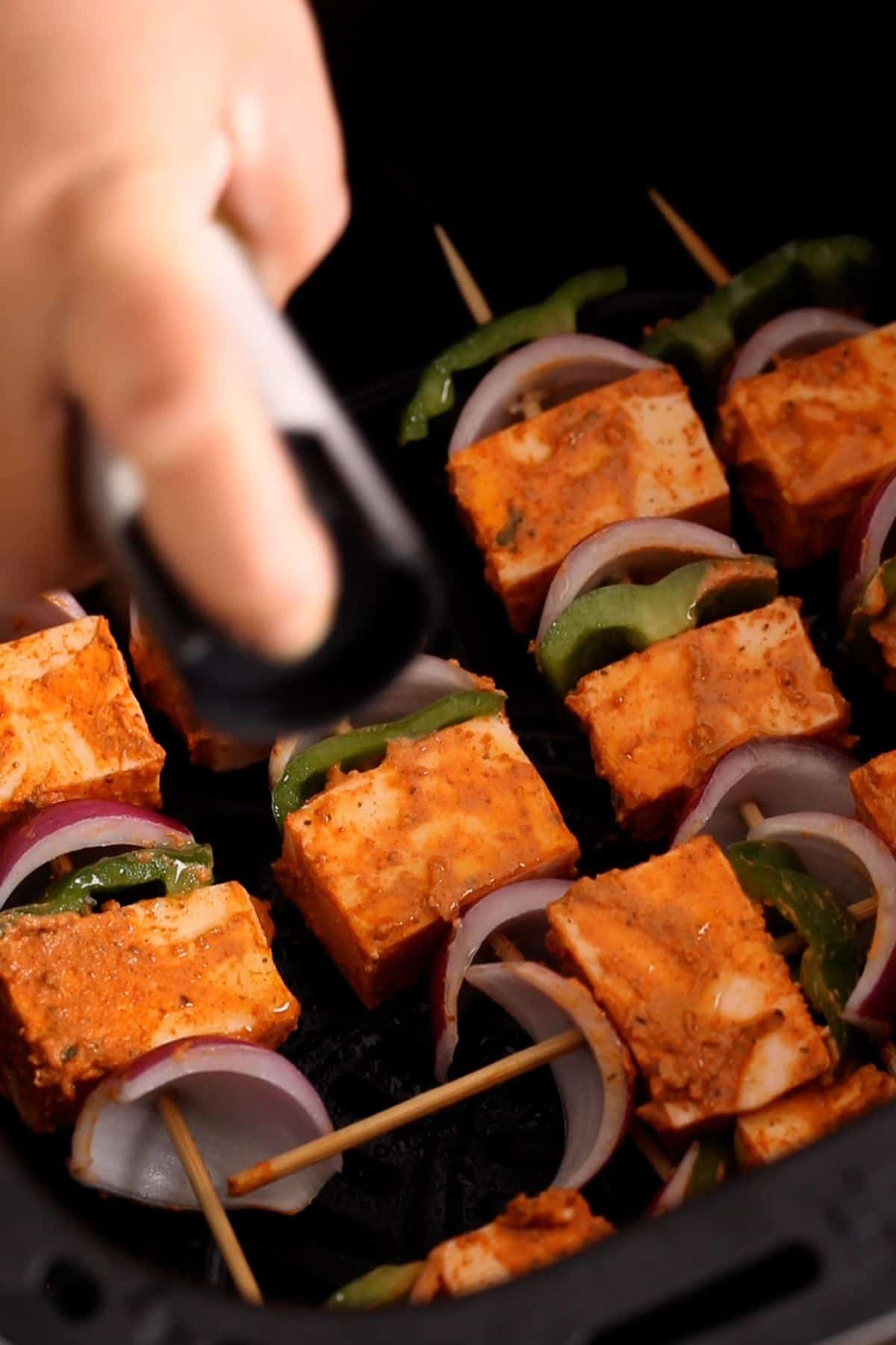 Paneer tikka skewers are placed in air fryer basket and sprayed with oil.