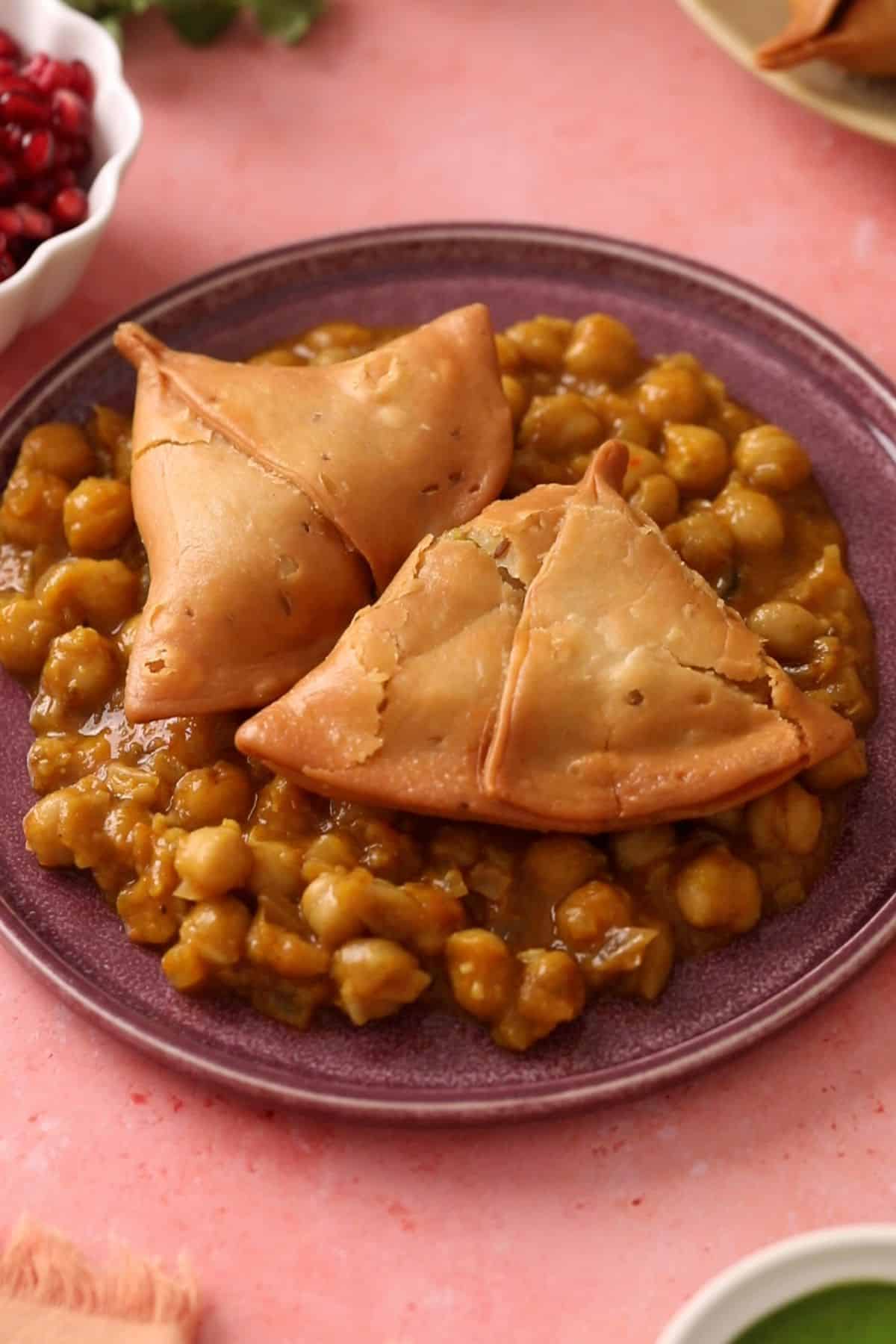Chickpeas curry and smashed samosa are placed on a purple plate.