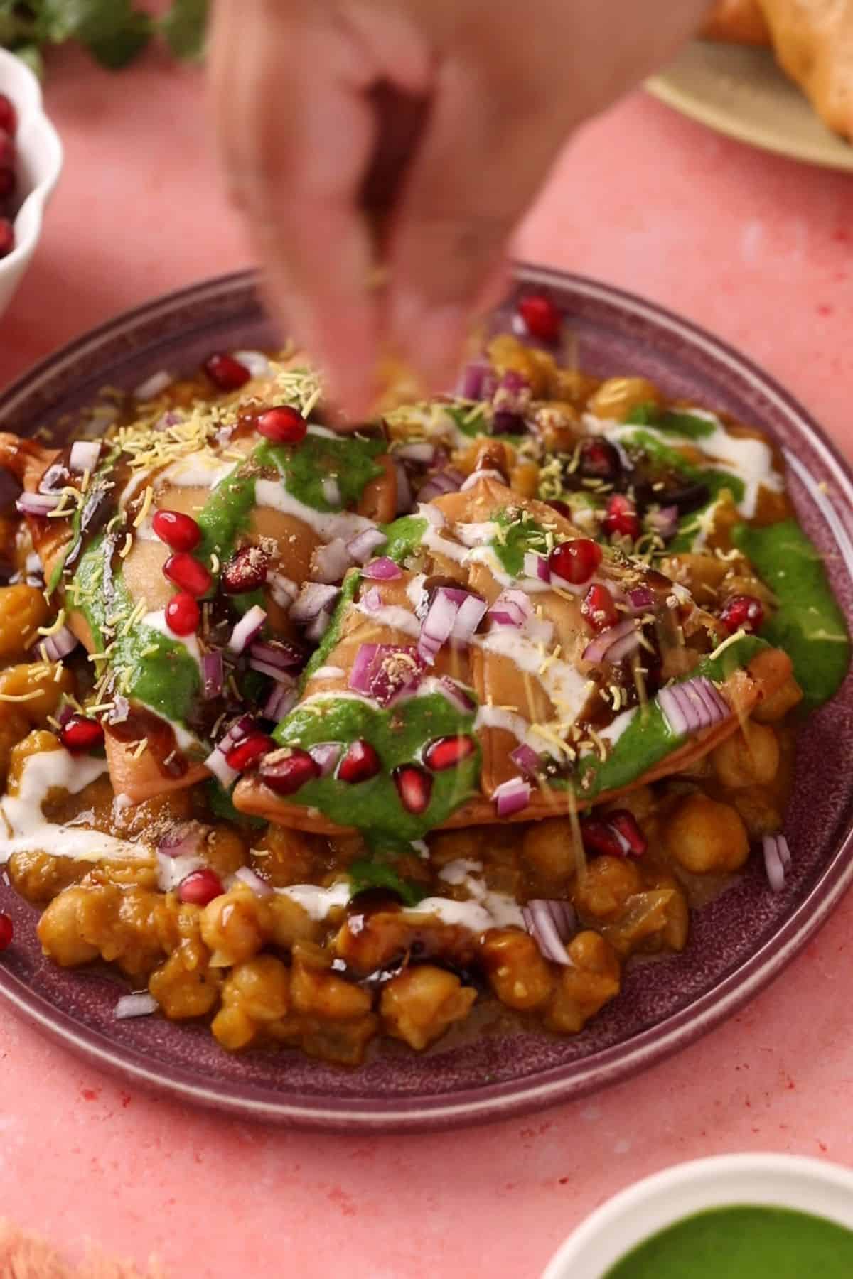 Samosa chaat is topped with onions, chaat spices, sev, pomegranate and cilantro.
