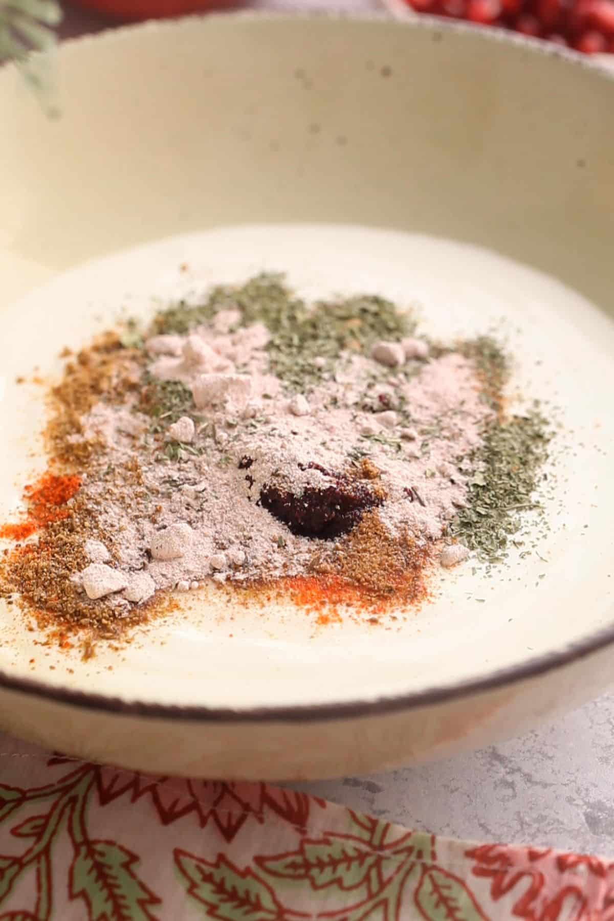 Spices, dried mint, salt and sugar are added to beatn yogurt in a bowl.