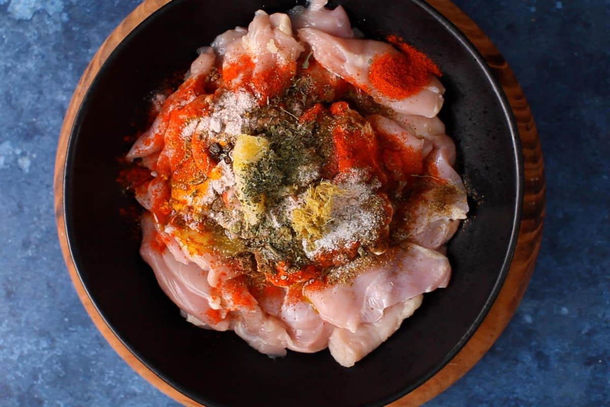 Raw chicken pieces in a black bowl are topped with ingredients of tandoori marinade.