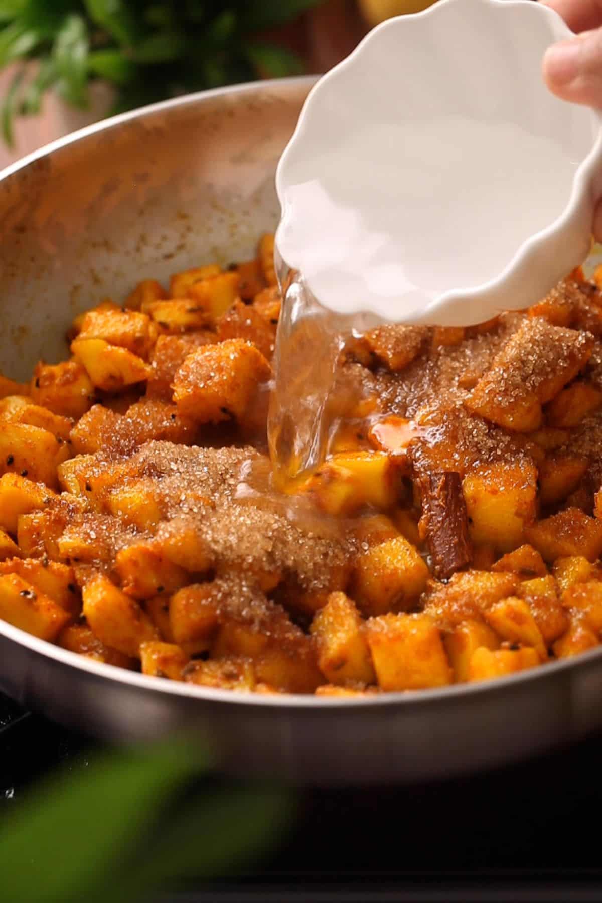 Close-up of a hand pouring water from a scalloped white dish onto a pan filled with diced mangoes, some coated in brown sugar and visible cinnamon sticks.