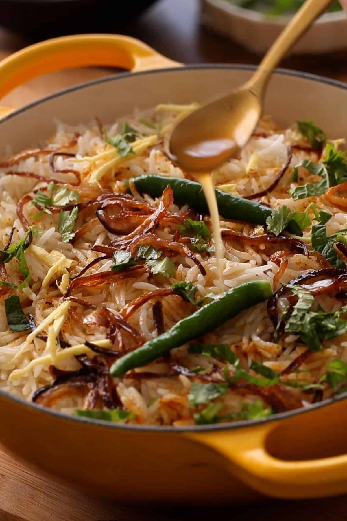 A close-up of biryani in a yellow pot topped with fried onions, green chilies, fresh cilantro, and ginger. Ghee is drizzled over the dish with a golden spoon.
