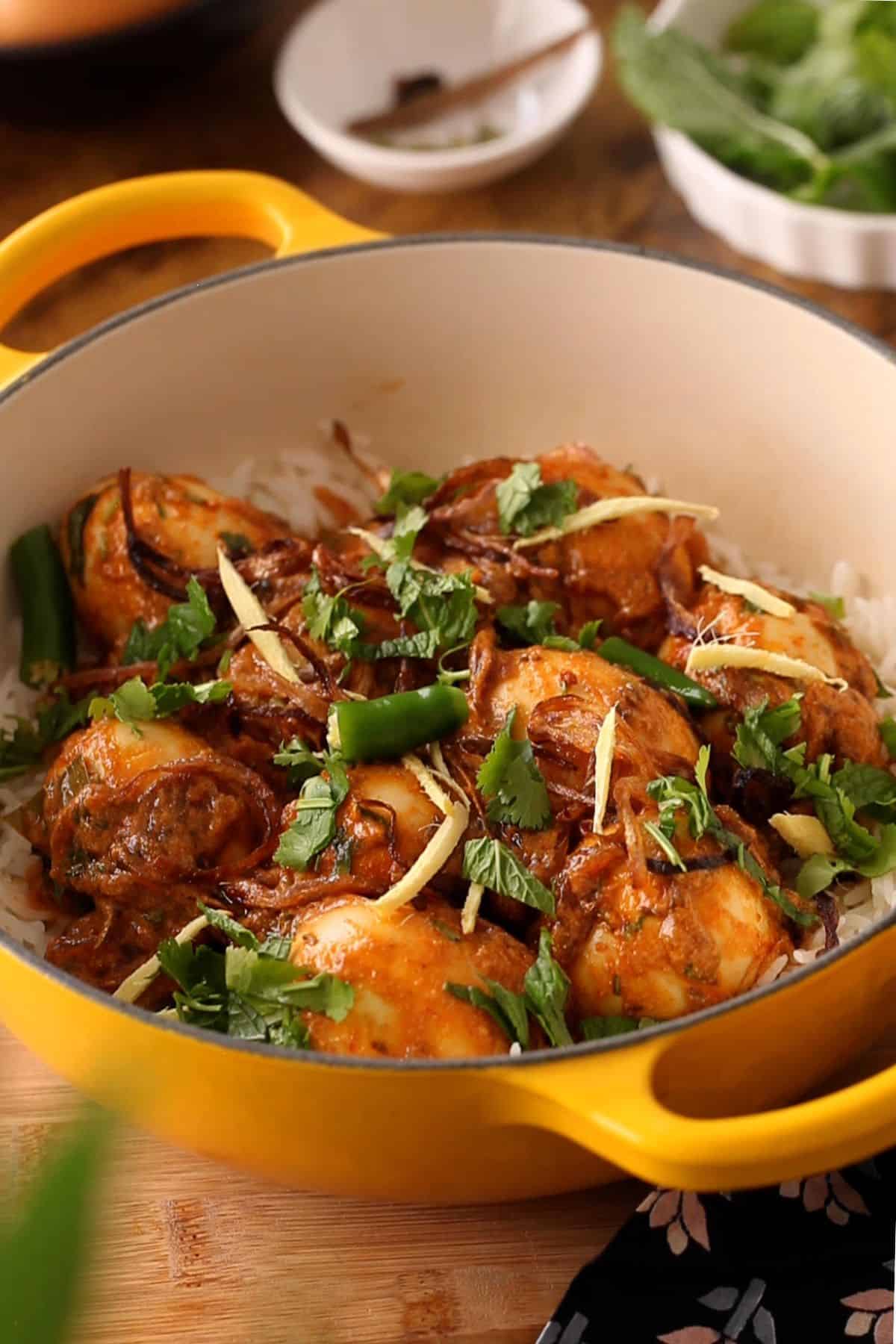 A yellow pot filled with spiced curry eggs garnished with fresh cilantro, sliced green chilies, and ginger strips, placed over a bed of rice.