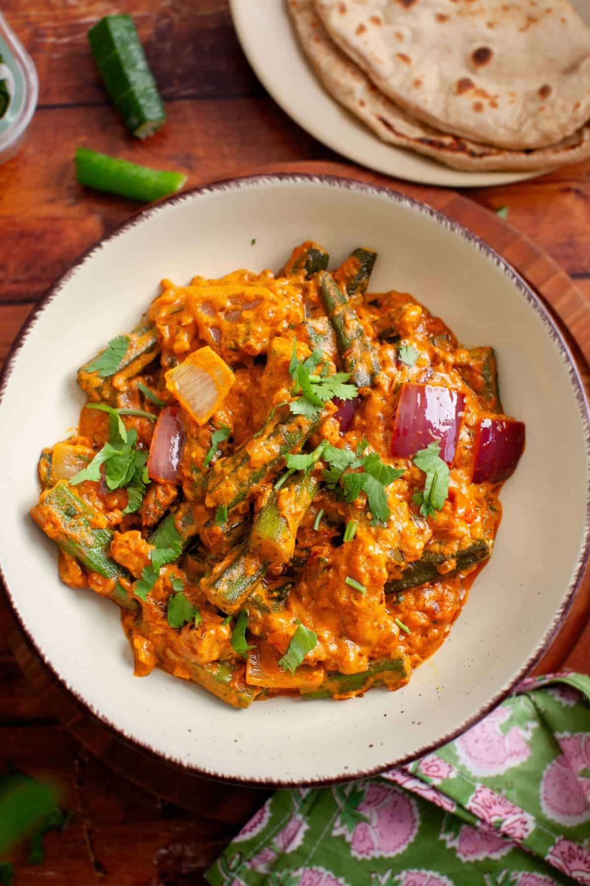 A bowl of bhindi masala sits on a wooden table with chapatis and a green chili in the background. 