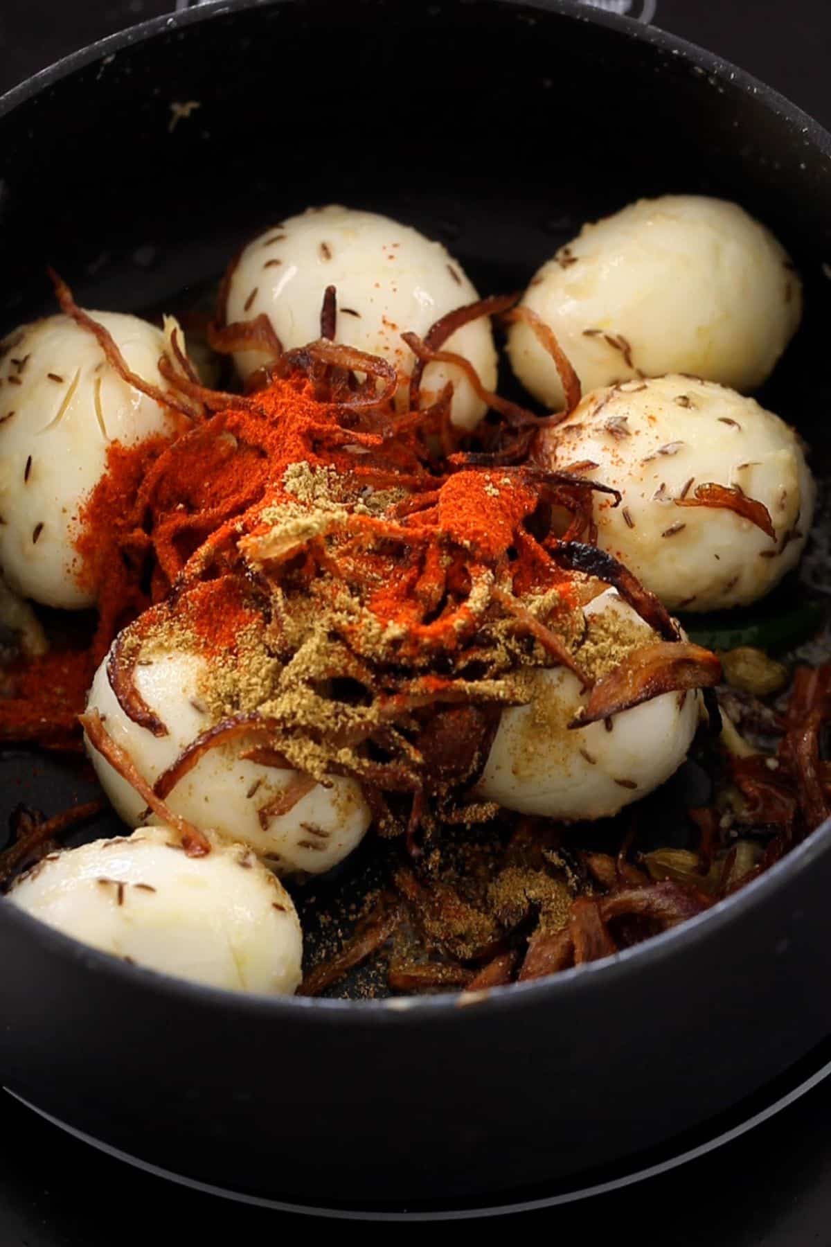 Eggs in a black cooking pot topped with fried onions and sprinkled with spices.