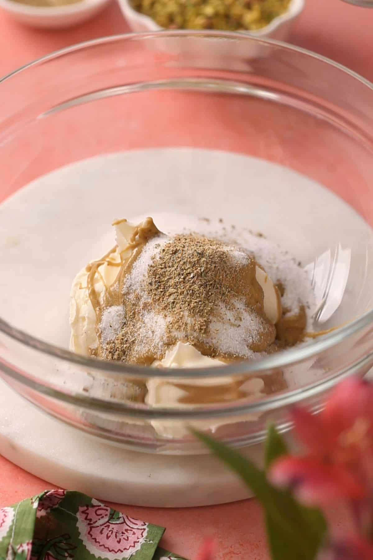A glass bowl containing cream, pistachios, soft cheese and spices sits on a marble surface.