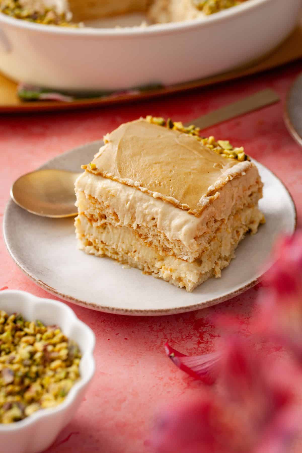 A slice of creamy layered tiramisu topped with crushed pistachios sits on a white plate with a gold spoon.