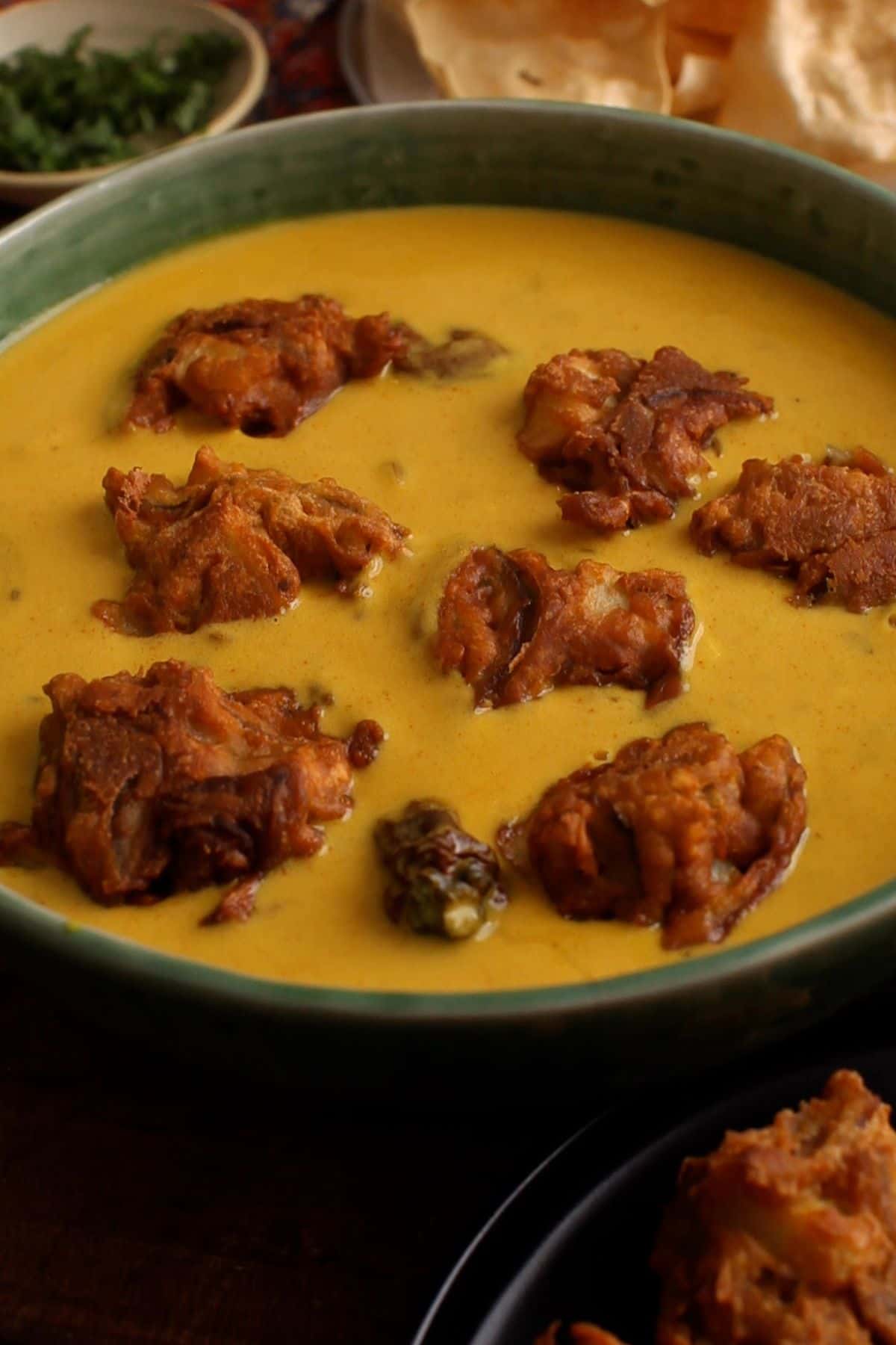 Fried pakoras are added to top of kadhi in a green bowl.