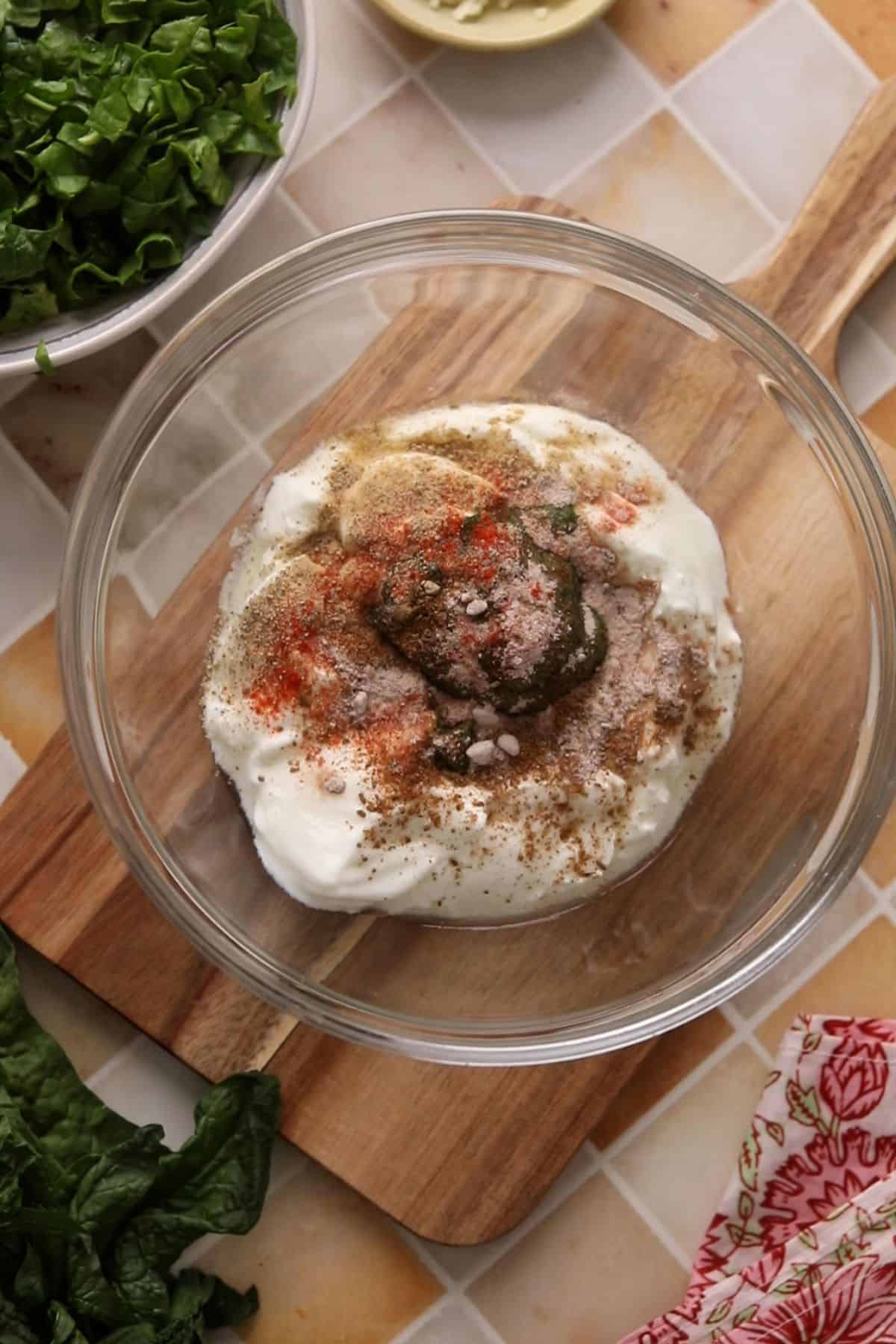 Yogurt, spinach puree and spices are added to a glass bowl.