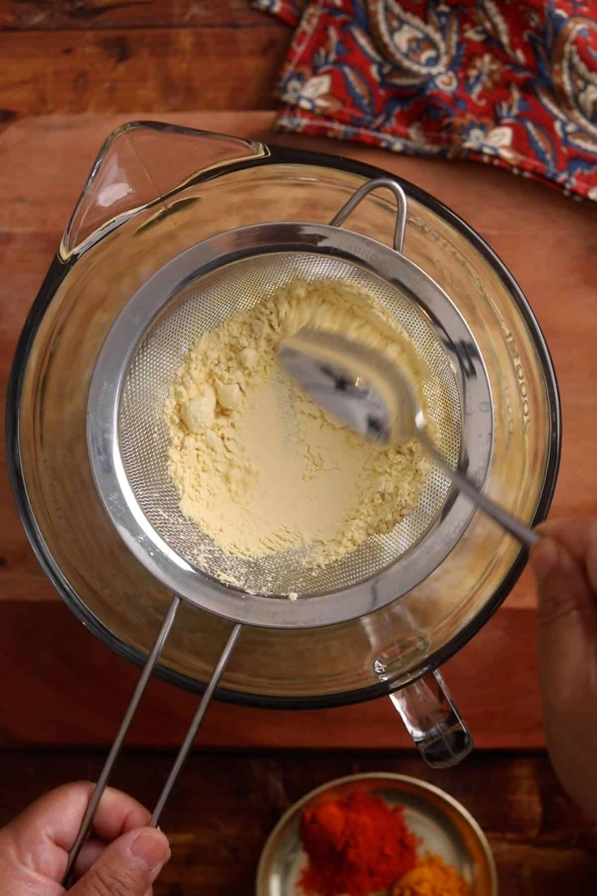 Sieving besan flour through stainless steel strainer over yogurt contained in a glass bowl.