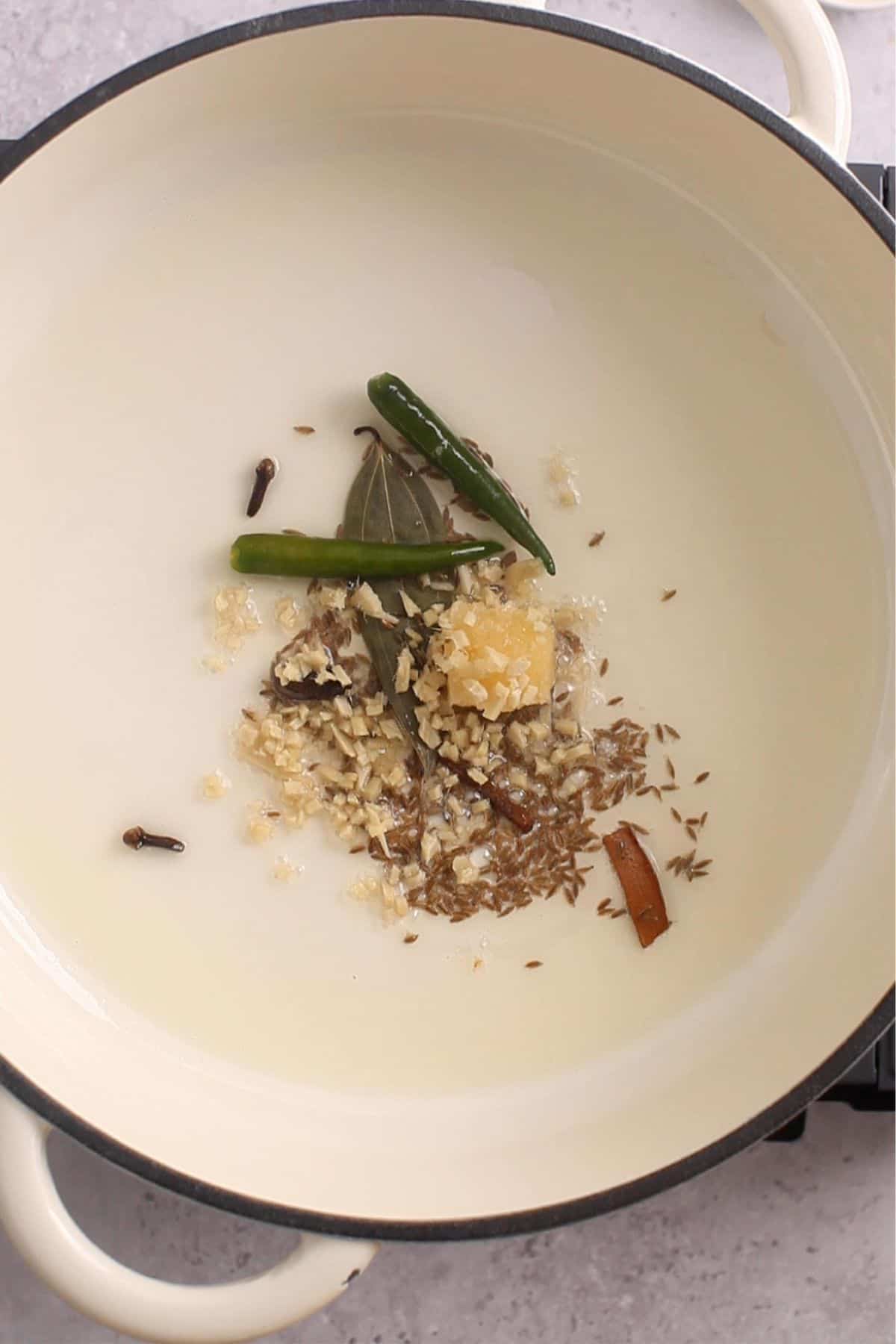 Whole spices & aromatics are added to hot oil in a white cooking pot.