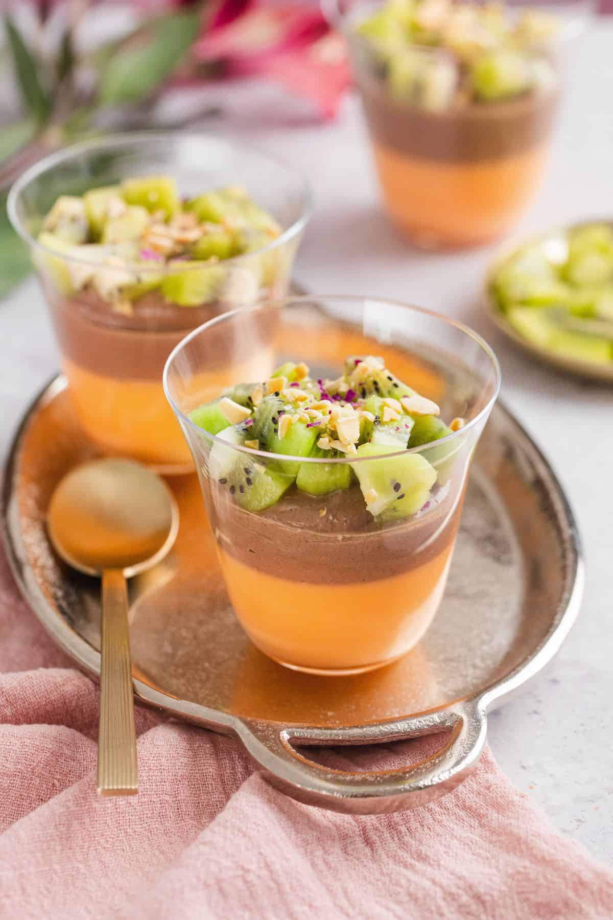A silver tray has two dessert cupos of orange jelly and chocolate shirkhand dessert with cut up kiwi on top.